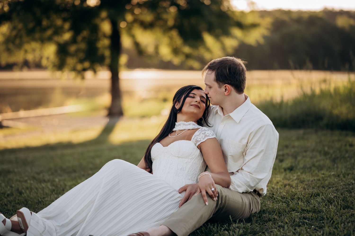 Couple sitting on grass by a lake at golden hour, woman in white dress leaning back smiling as man whispers to her.