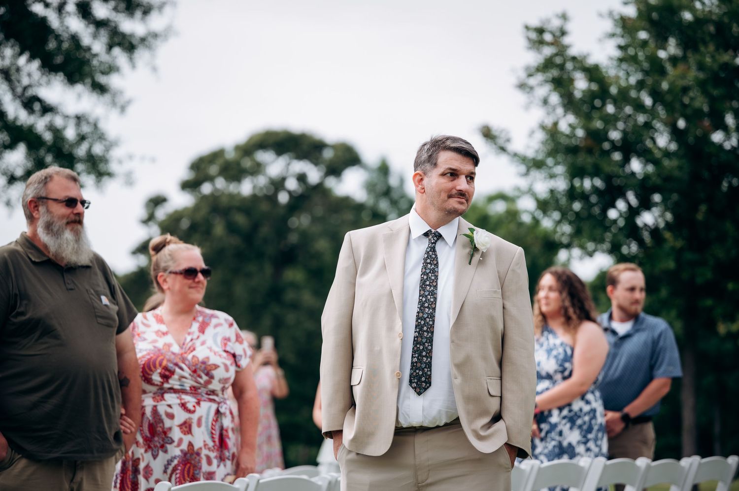 Smiling man in tan suit walks down the aisle at an outdoor wedding ceremony among seated guests.