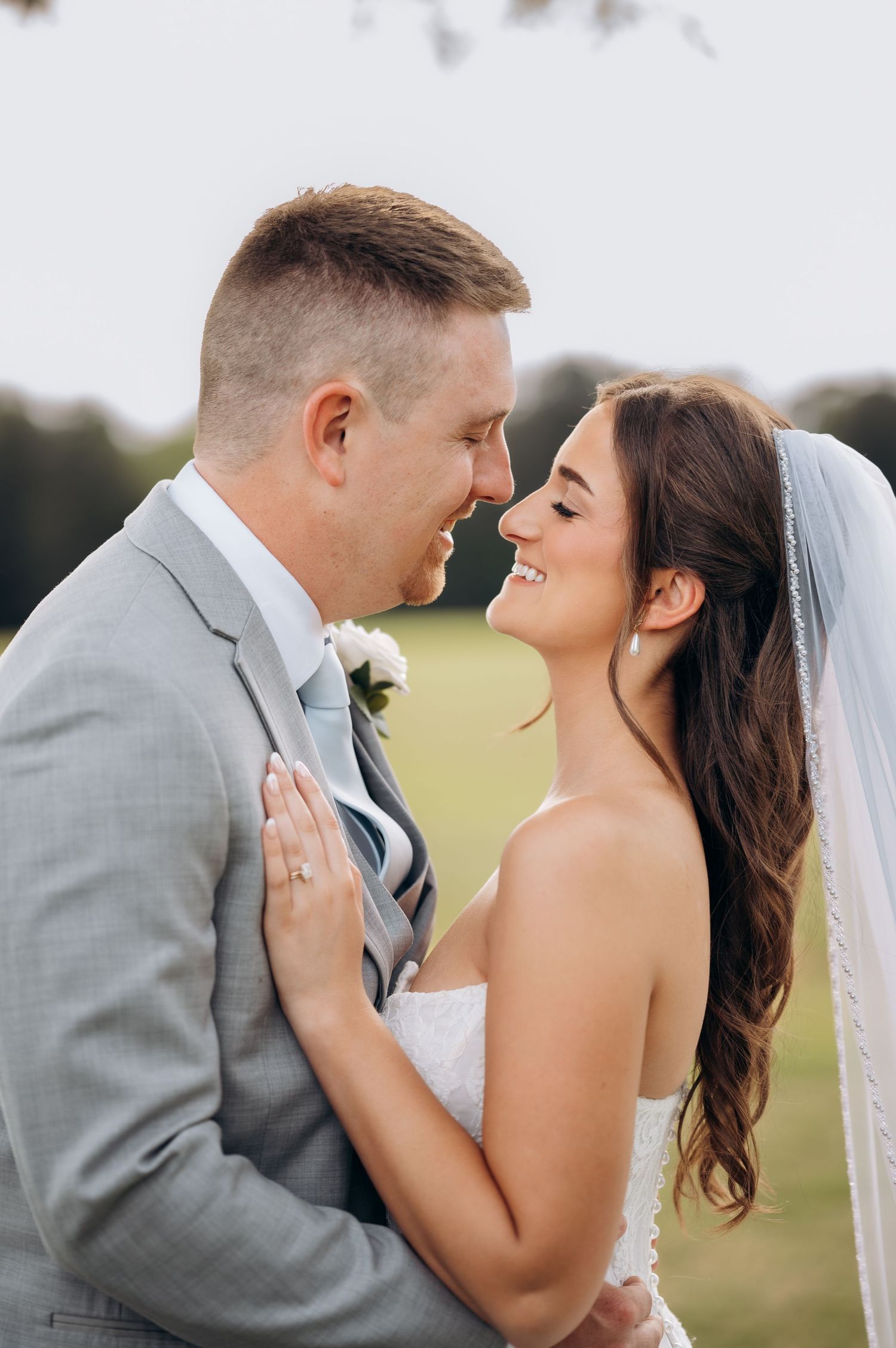 Smiling bride and groom touching foreheads outdoors, groom in gray suit, bride in white strapless gown with veil.