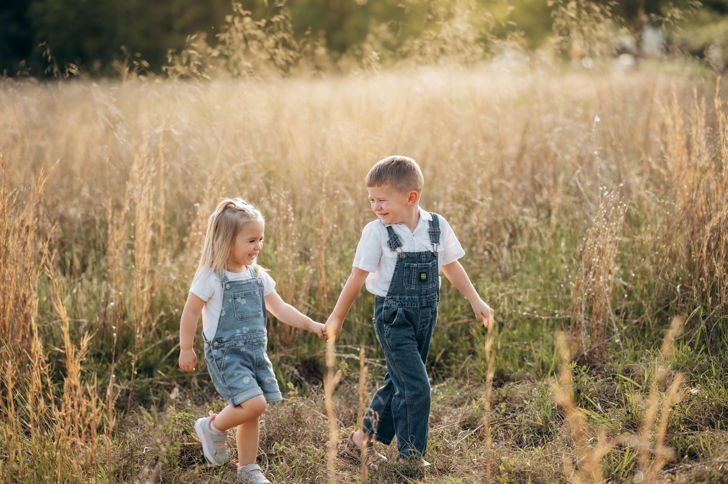 Two young children in denim overalls hold hands while walking through a sunlit golden field.