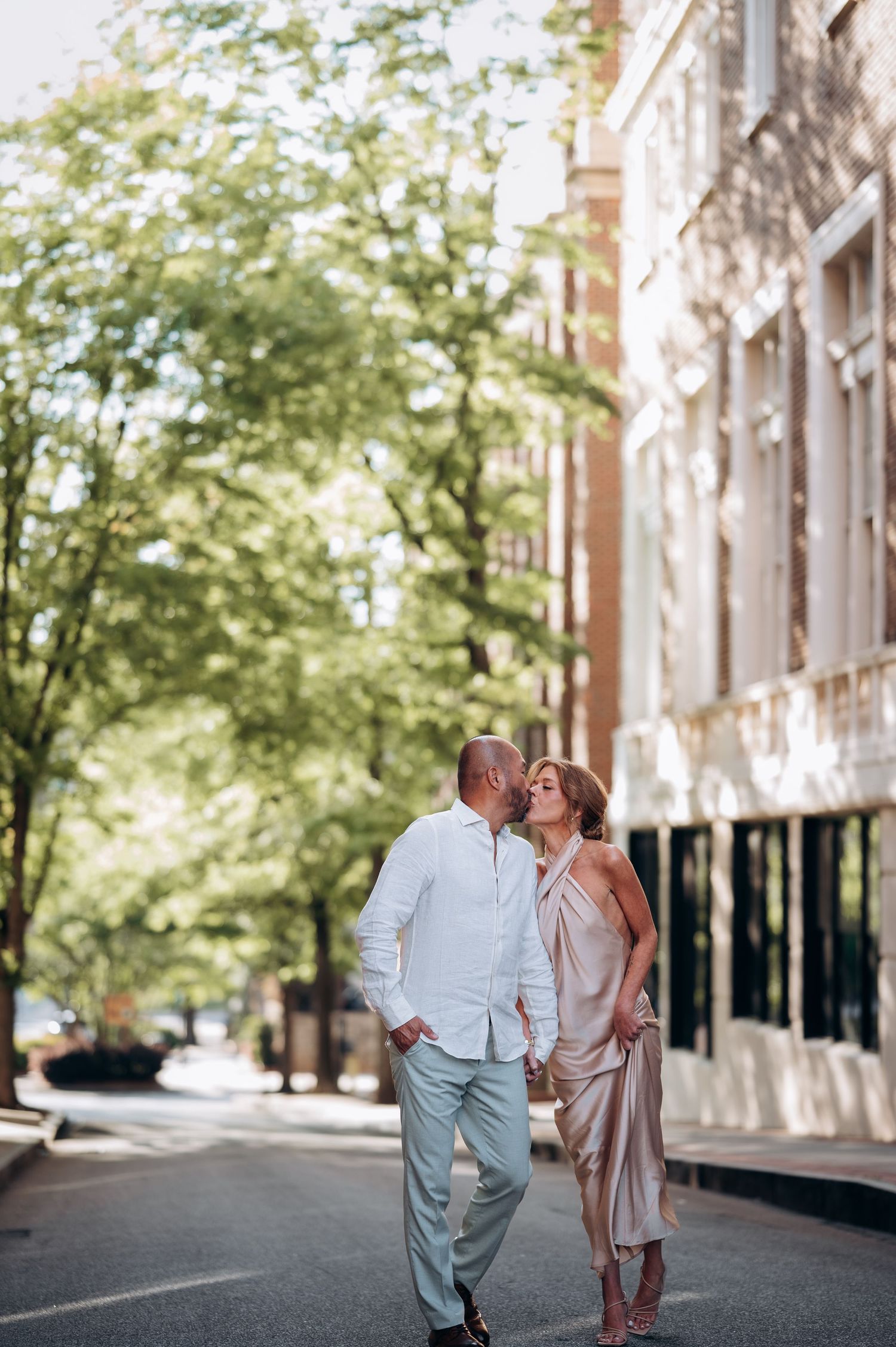 Couple sharing a kiss while strolling on a tree-lined city street, dressed in elegant casual attire.