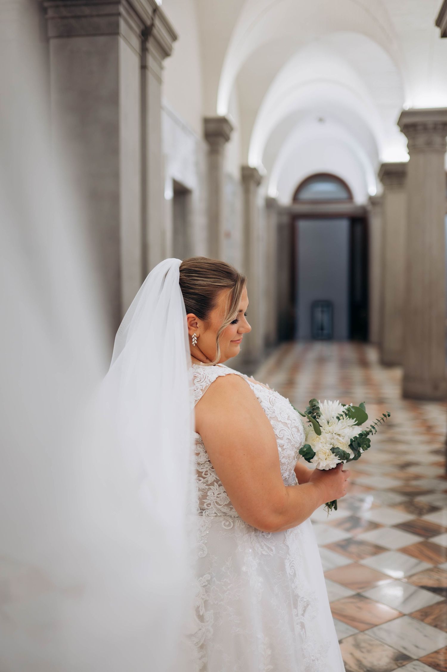 Bride holding bouquet walks through grand hallway with arched ceilings and checkered floor.