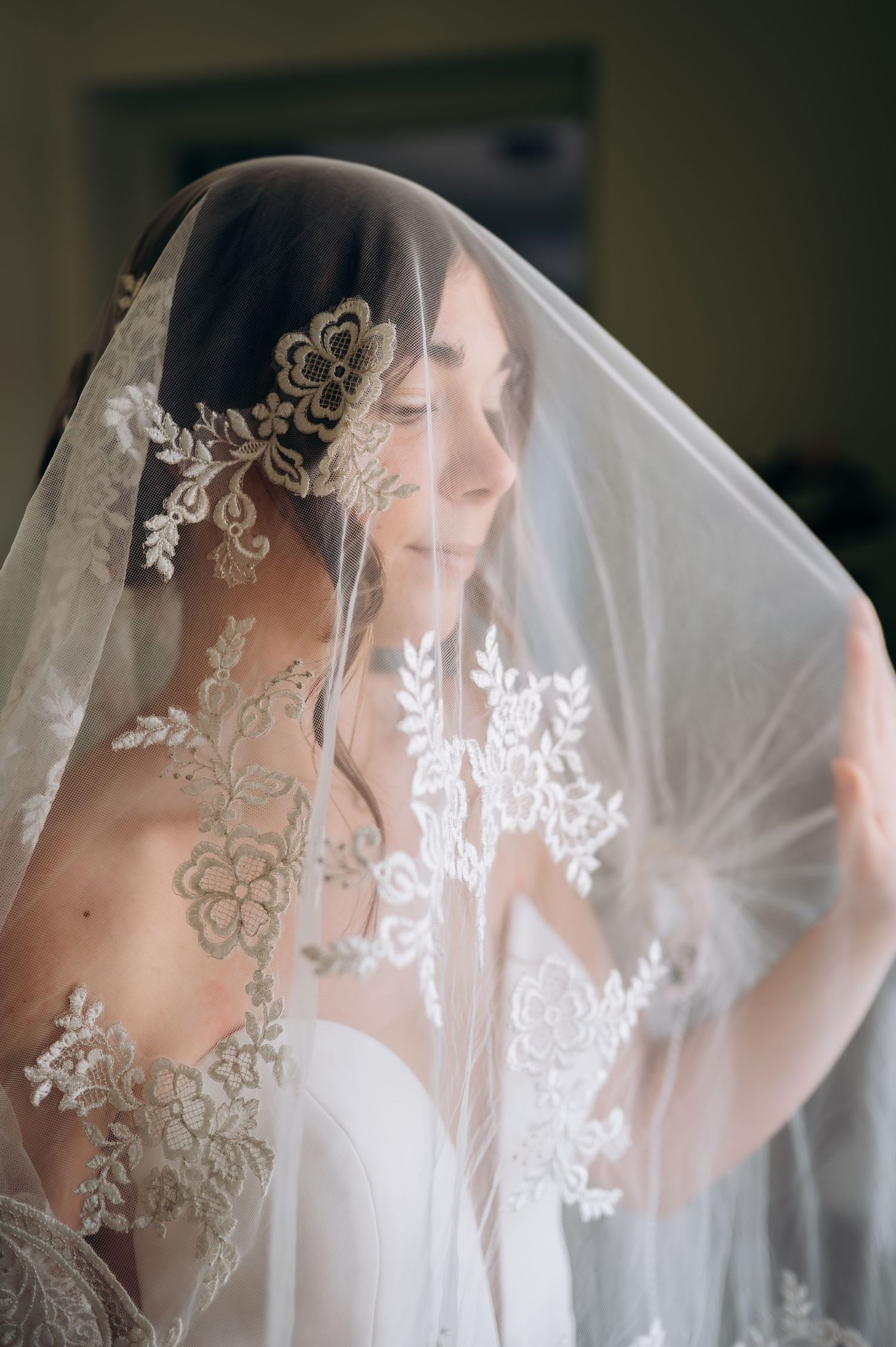 Bride draped in an elegant lace-trimmed veil with floral embroidery smiling softly on wedding day.