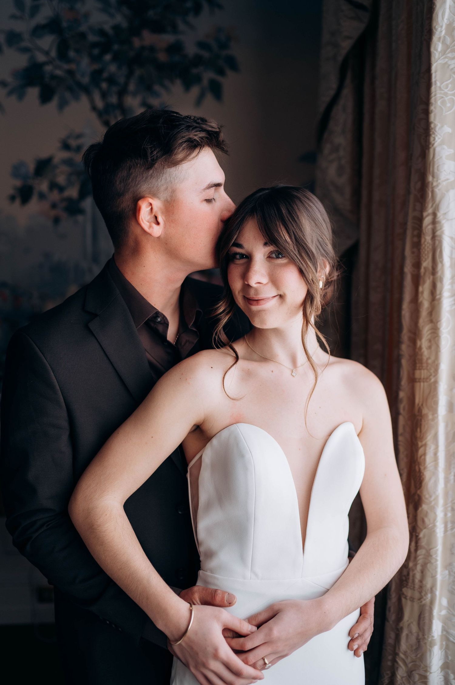 Groom in black suit kisses bride on cheek as she smiles warmly in strapless white wedding gown indoors.