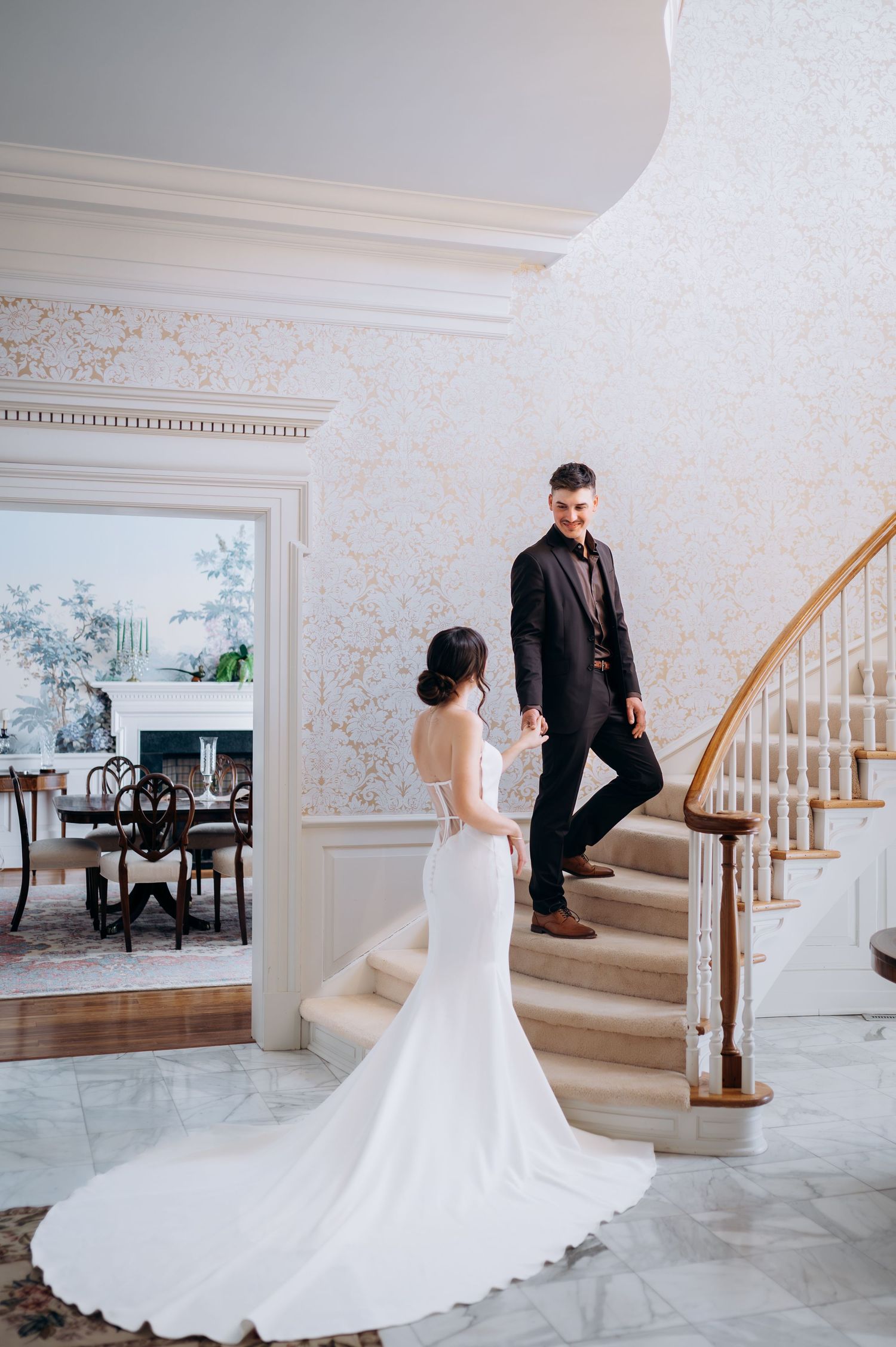 Bride in backless white gown holds grooms hand on a grand curved staircase in an elegant estate.