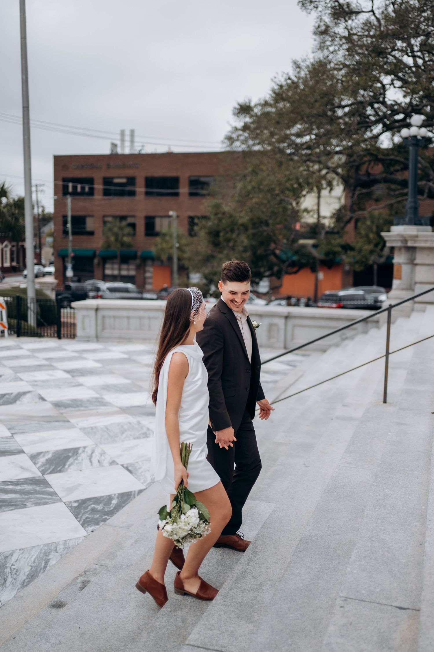Newlyweds walk hand-in-hand up marble steps, bride holding white bouquet in short white dress.