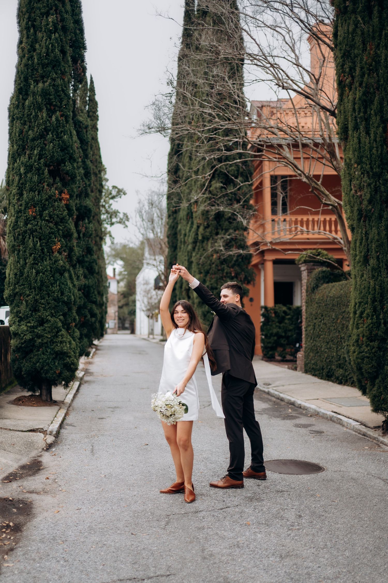 Groom twirling bride in white mini dress on a tree-lined street during their wedding day celebration.