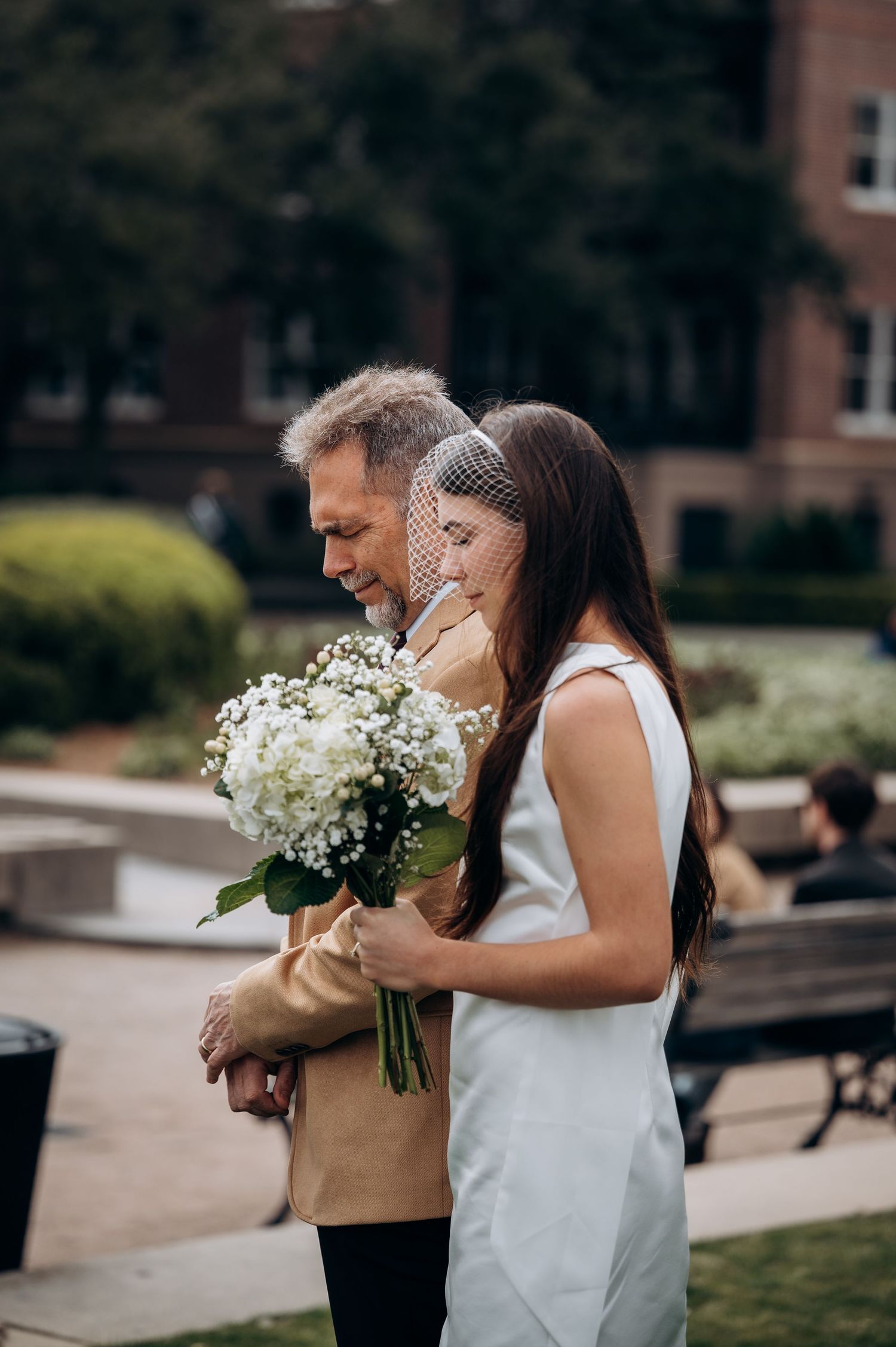 Bride in white dress and veil embraces groom outdoors holding white hydrangea bouquet at wedding ceremony.