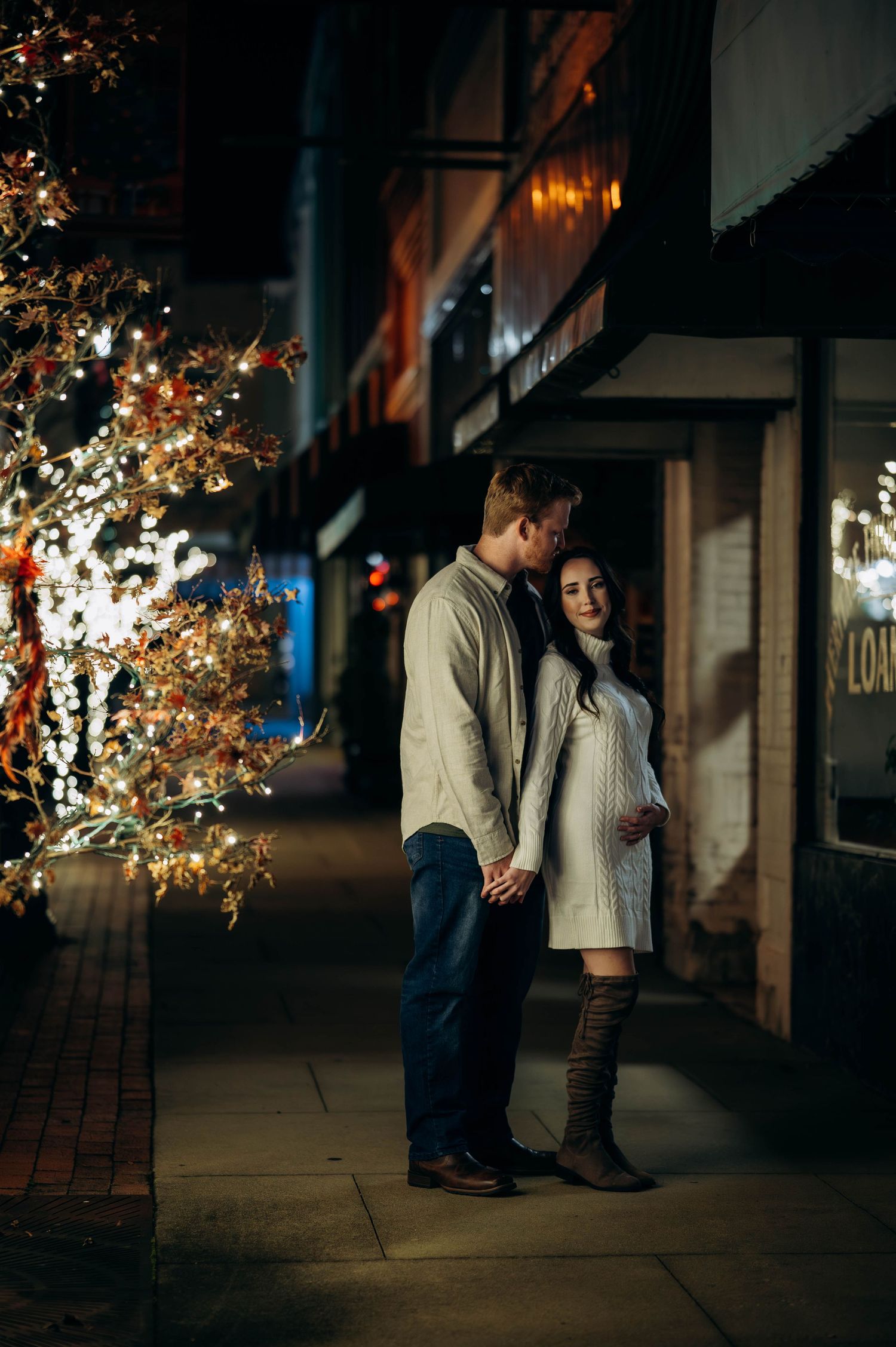 Expecting couple sharing intimate moment on decorated night street, man tenderly looking at pregnant partner in white dress.