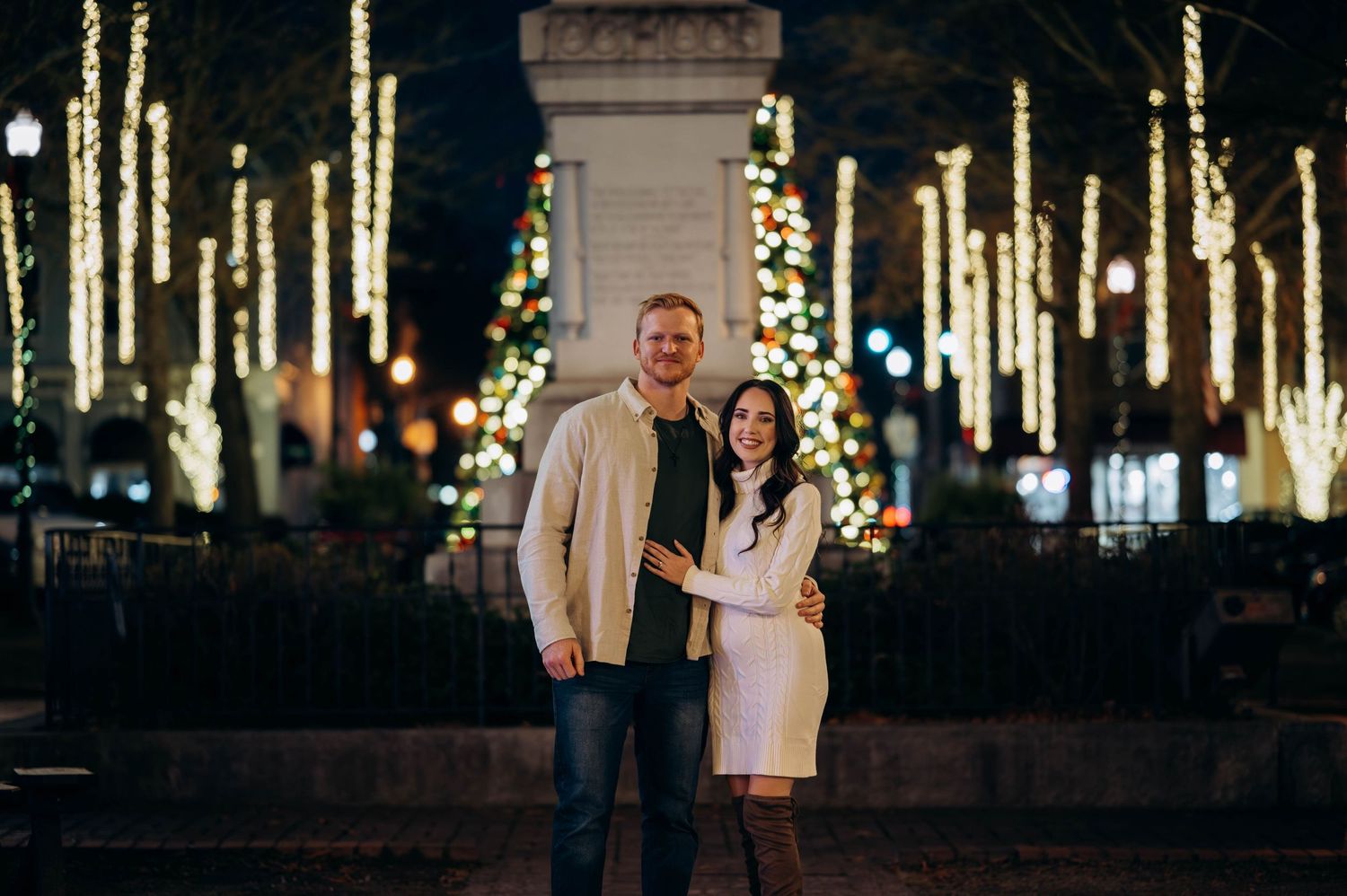 Couple smiling together in front of festive Christmas lights and decorated tree at night.