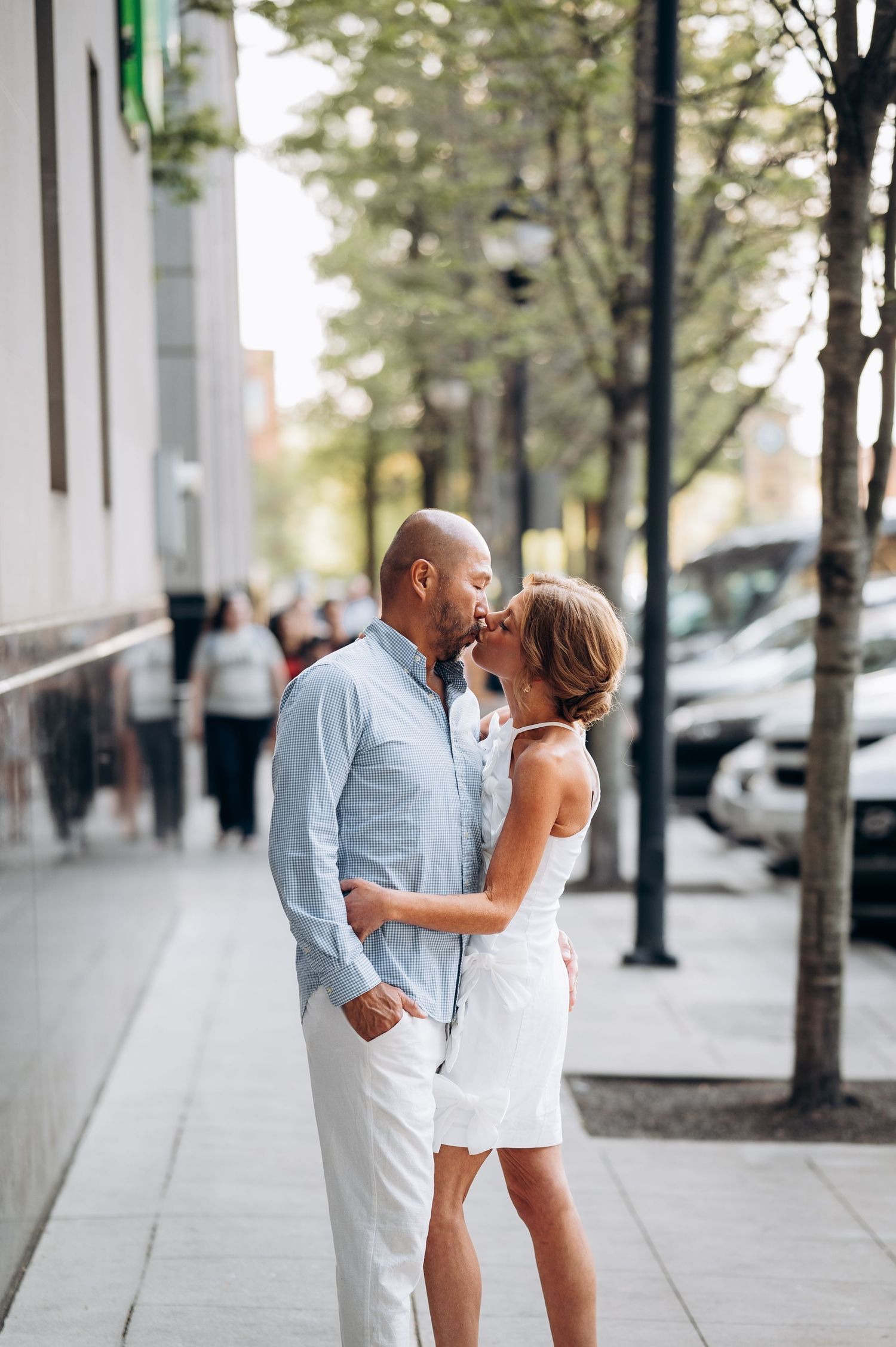 Loving couple almost kissing on a sunny urban sidewalk lined with trees and parked cars.