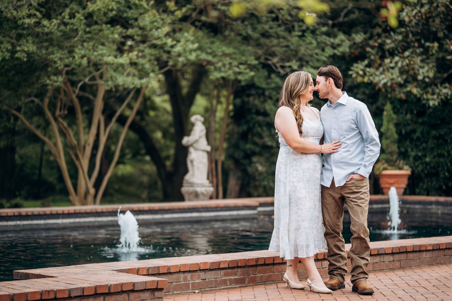 Romantic couple embracing near an elegant fountain with classical statue in a formal garden.