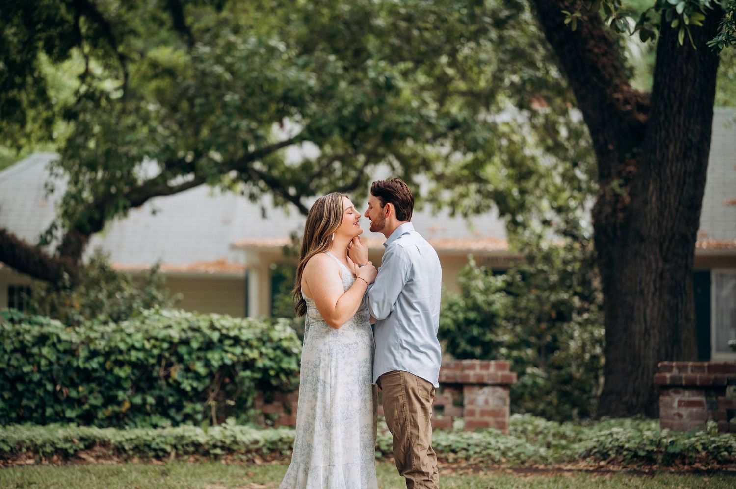 Couple sharing a kiss on a green lawn with large oak trees in the background.