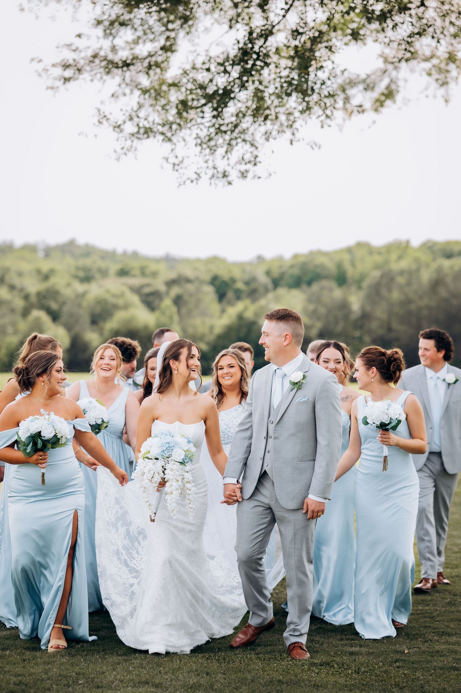 Bride and groom walk with wedding party in light blue dresses and gray suits on green lawn.