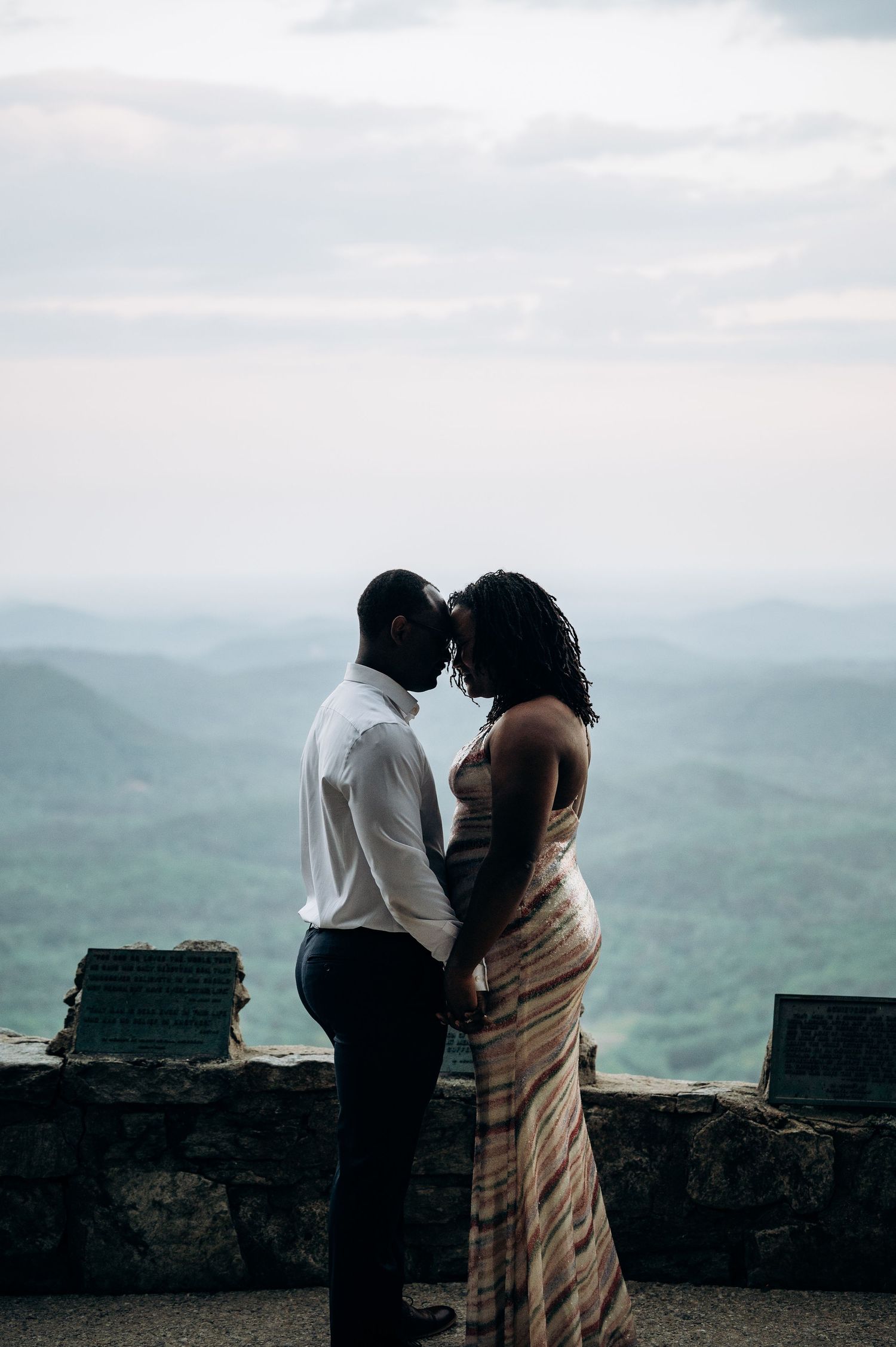 A couple shares an intimate moment on a mountain overlook, forehead to forehead, with lush green hills in the background.