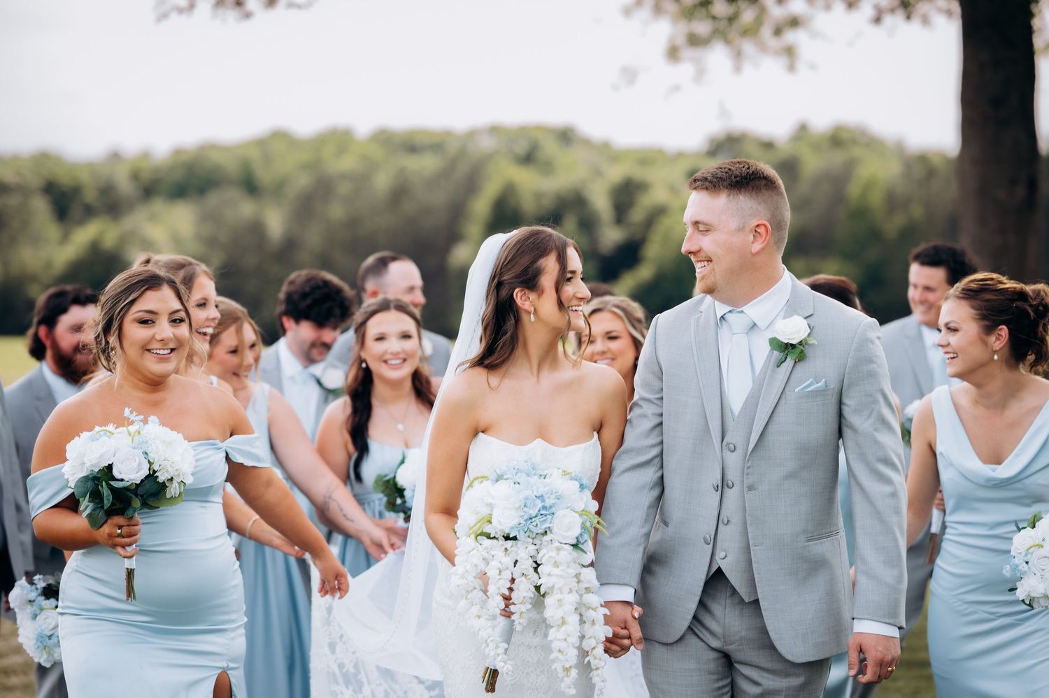 Wedding party walks outdoors, bride in white gown with cascading bouquet, groom in gray suit, bridesmaids in light blue dresses.