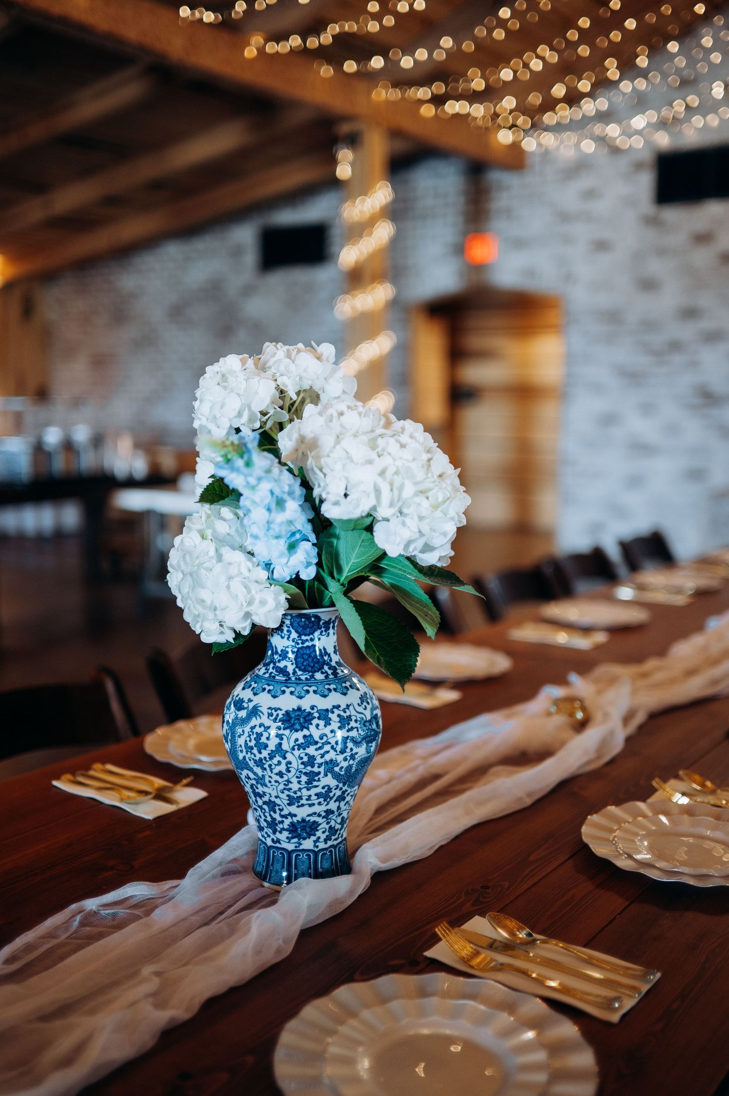 Blue and white floral vase with white hydrangeas on a rustic wooden table with gold cutlery and string lights.