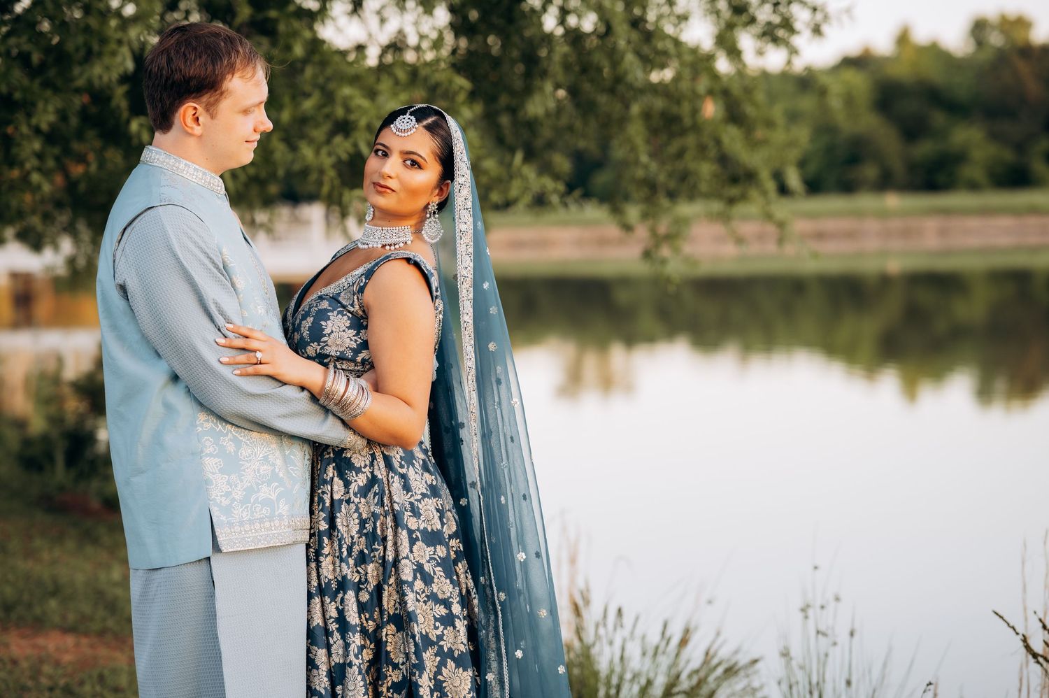 Couple in elegant blue attire poses by a serene lake, blending Western and South Asian wedding styles.
