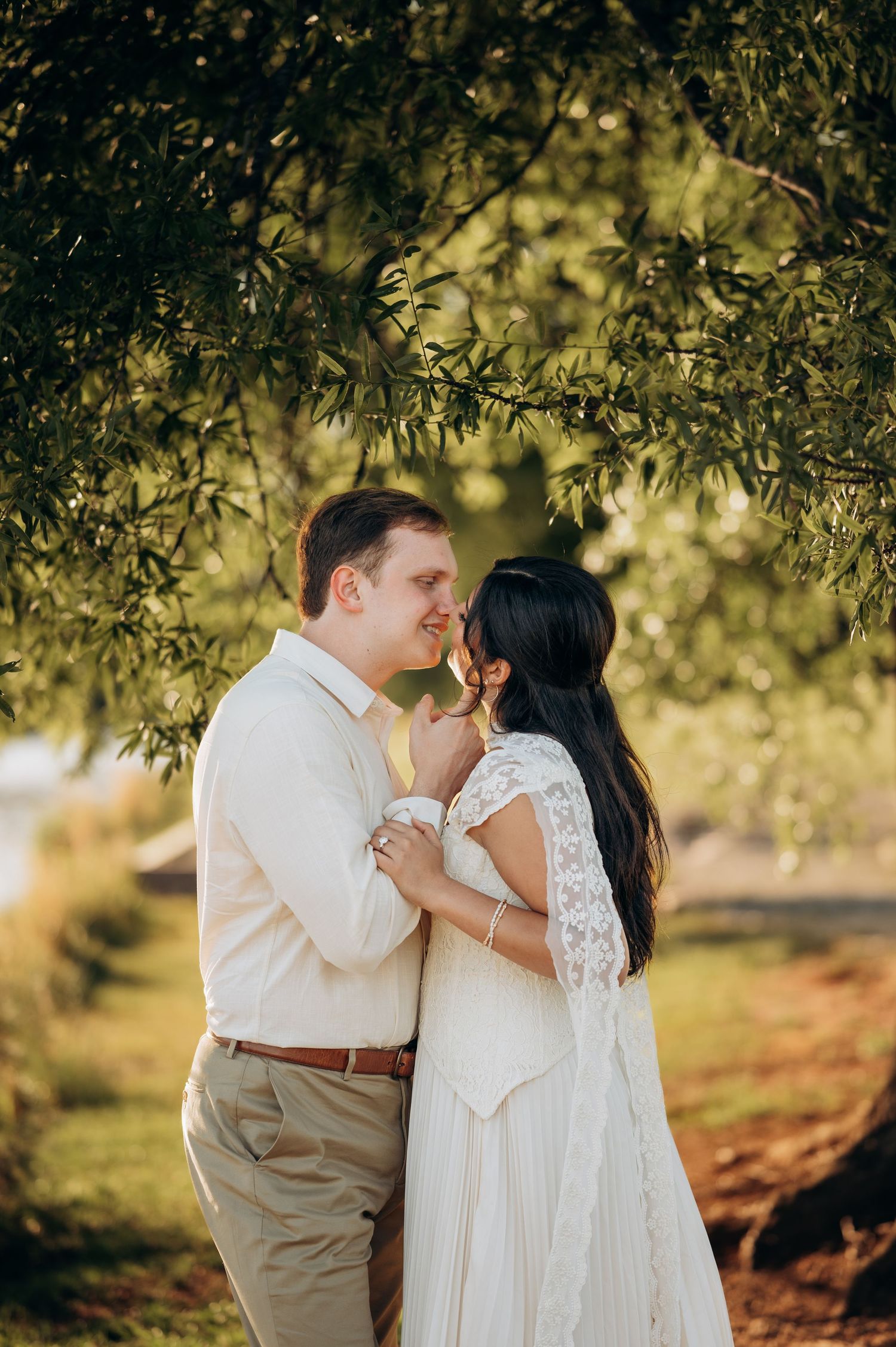 A couple shares a romantic moment under lush green tree branches, bride in white lace dress, groom in beige attire.