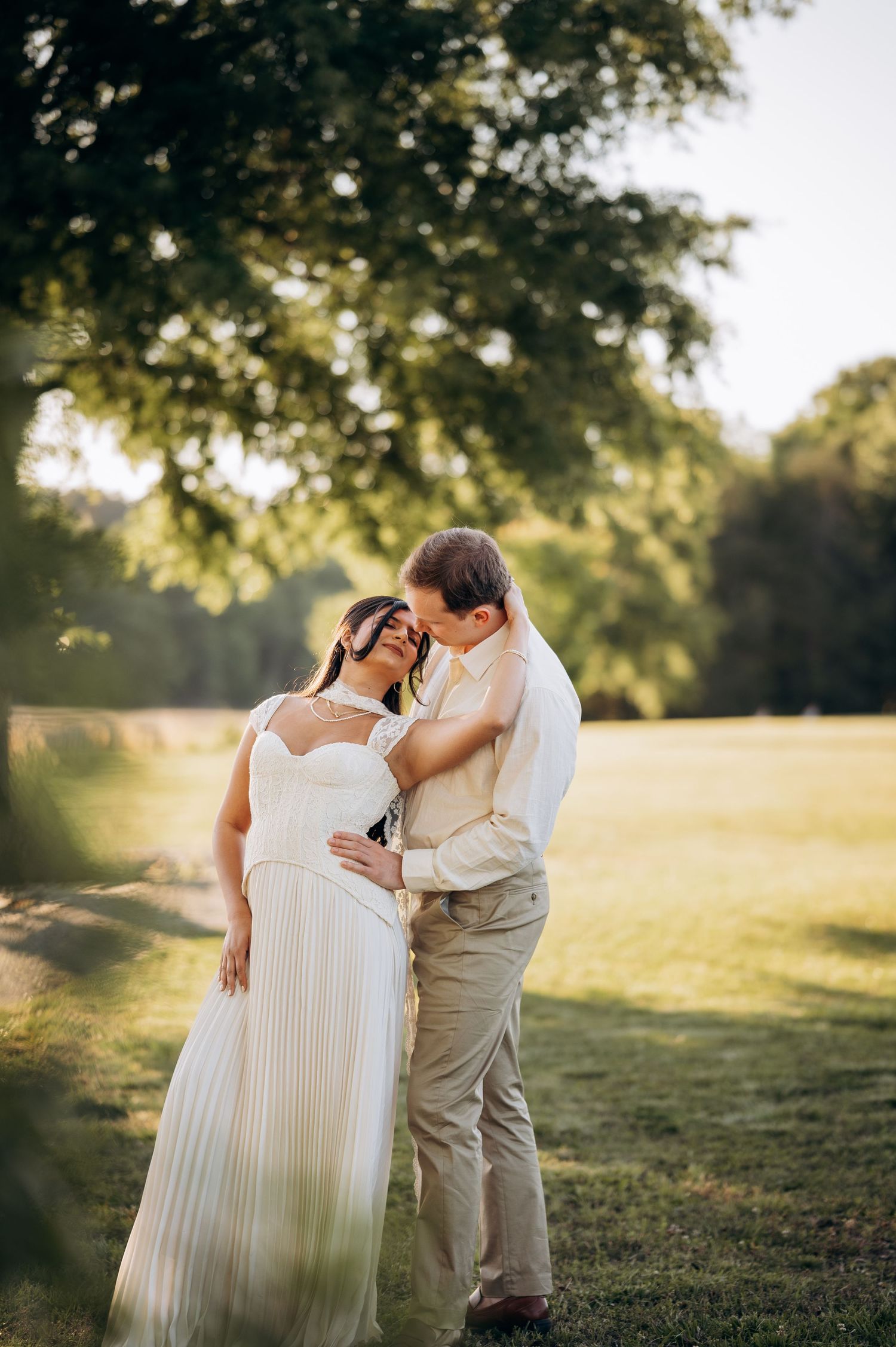 Couple sharing a romantic moment in a sunlit garden, bride in white pleated gown, groom in khaki attire.