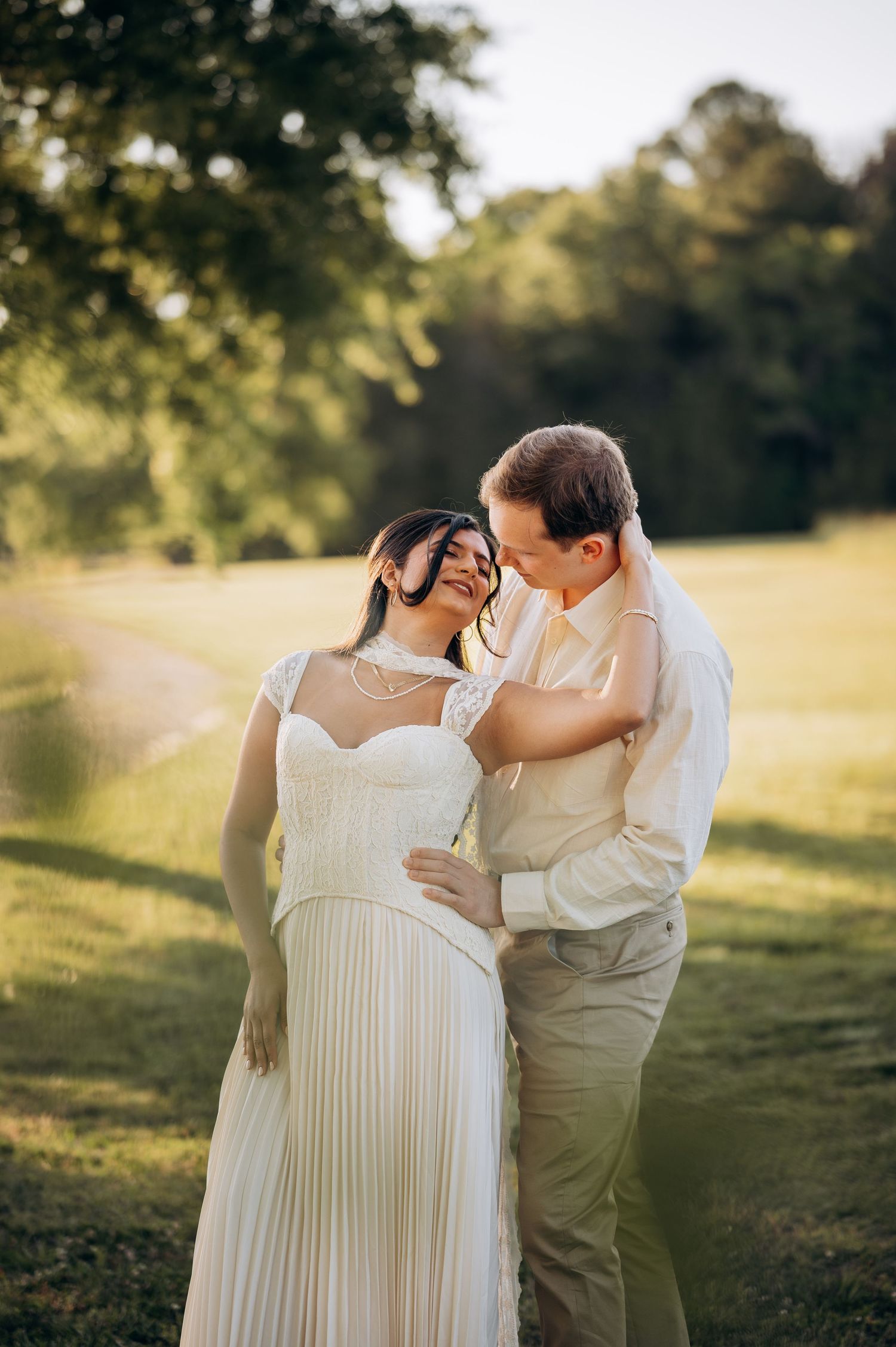 A couple shares a romantic moment outdoors, bride in white pleated gown with pearl necklace, groom in beige suit, golden hour light.