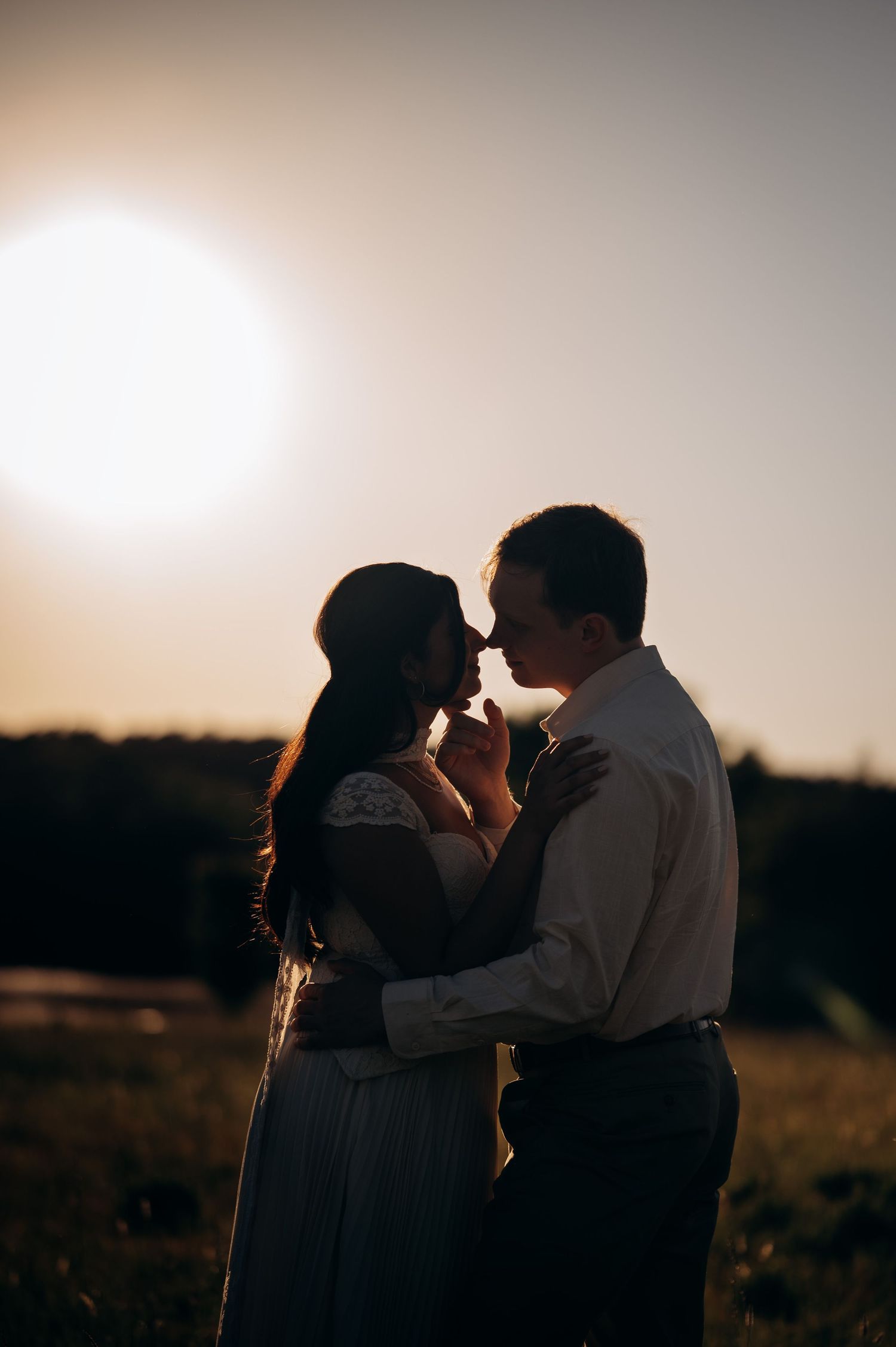 A couple shares a romantic moment at sunset in a field, silhouetted against the golden sky in wedding attire.