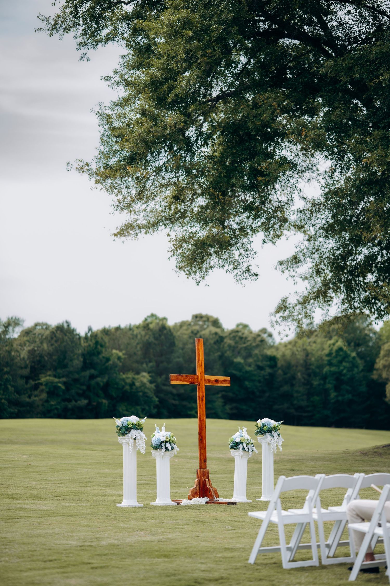 Outdoor wedding ceremony setup with wooden cross, white pillars, floral arrangements, and white chairs on a green lawn.