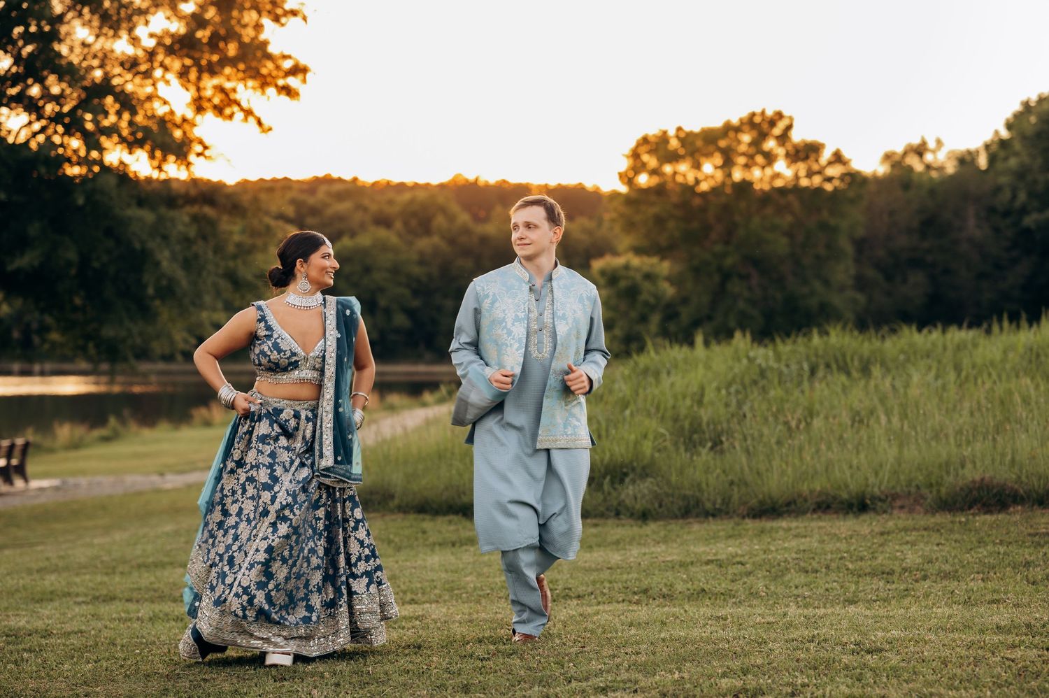Couple in Indian wedding attire walks outdoors at sunset, woman in blue lehenga, man in light blue sherwani, lush greenery behind.