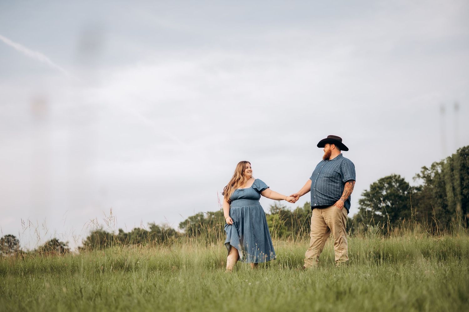 Couple holding hands in a green meadow, woman in blue dress, man in cowboy hat, walking together under cloudy sky.