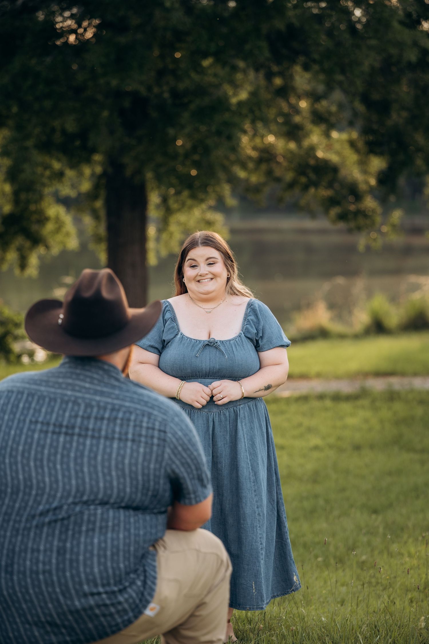 Woman in blue dress smiling joyfully as man in cowboy hat kneels before her near a lake.