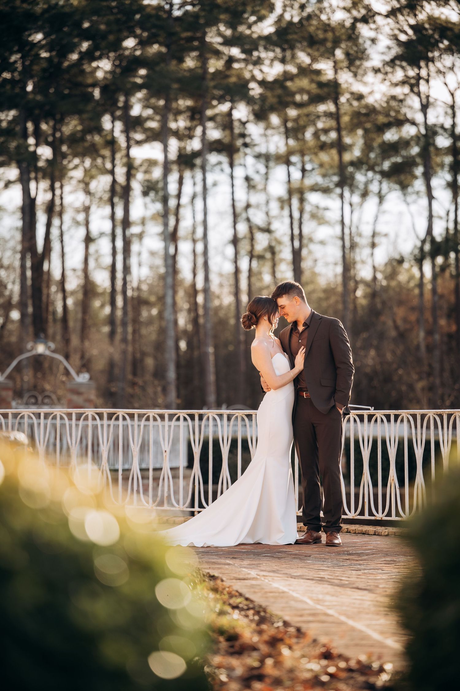 A romantic sunset wedding photo with a couple on a white bridge surrounded by pine trees.
