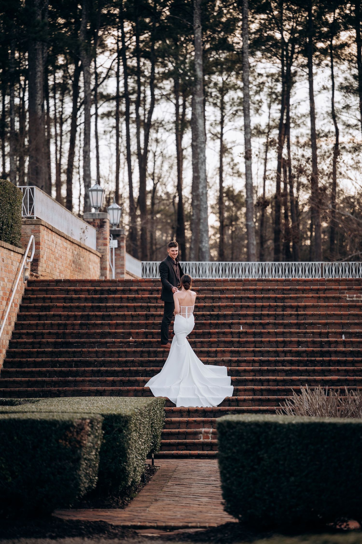 Bride posing on grand brick stairs surrounded by tall pine trees.