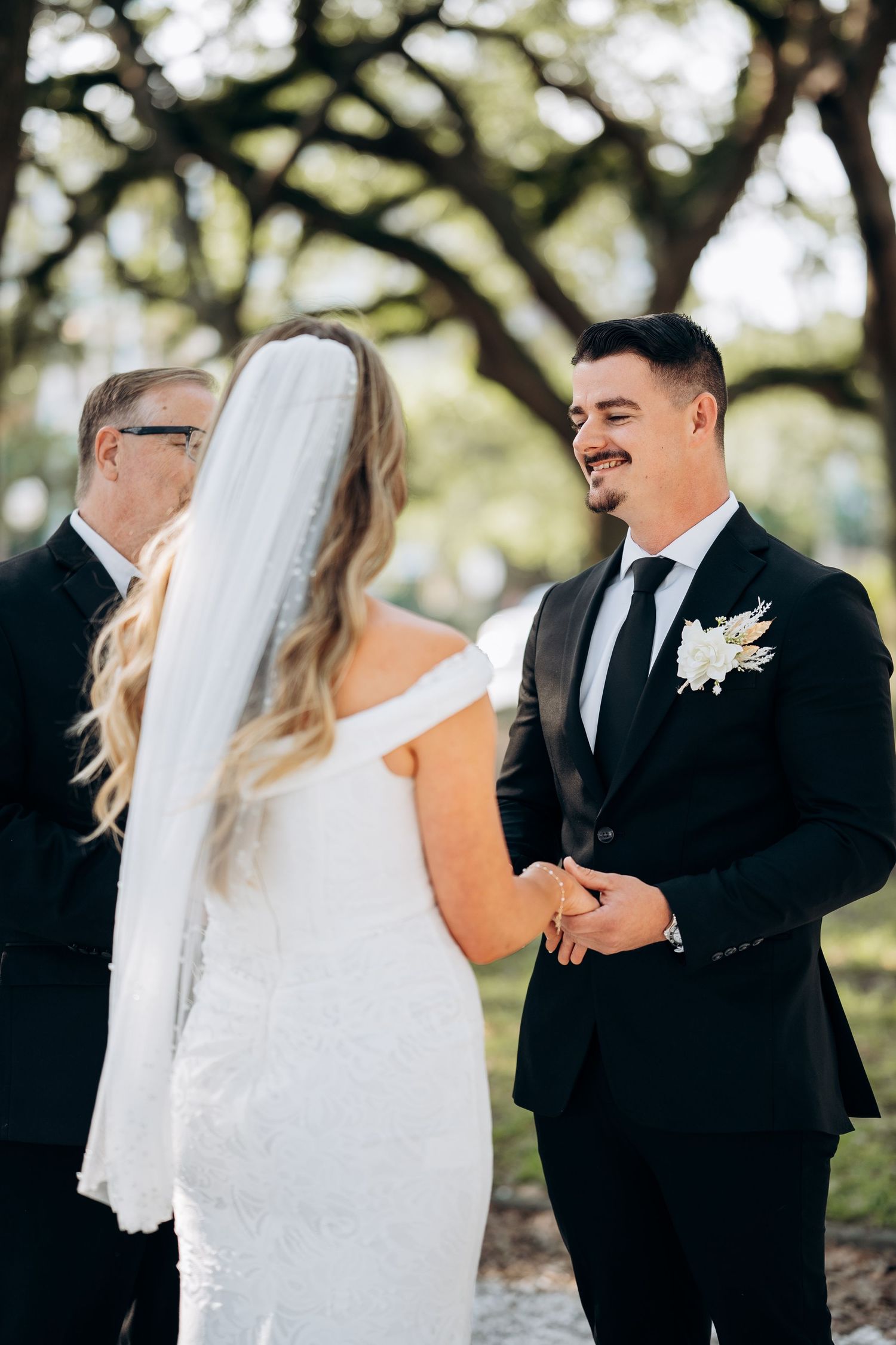 Groom smiling at bride during outdoor wedding ceremony under oak trees, wearing black suit with white boutonniere.