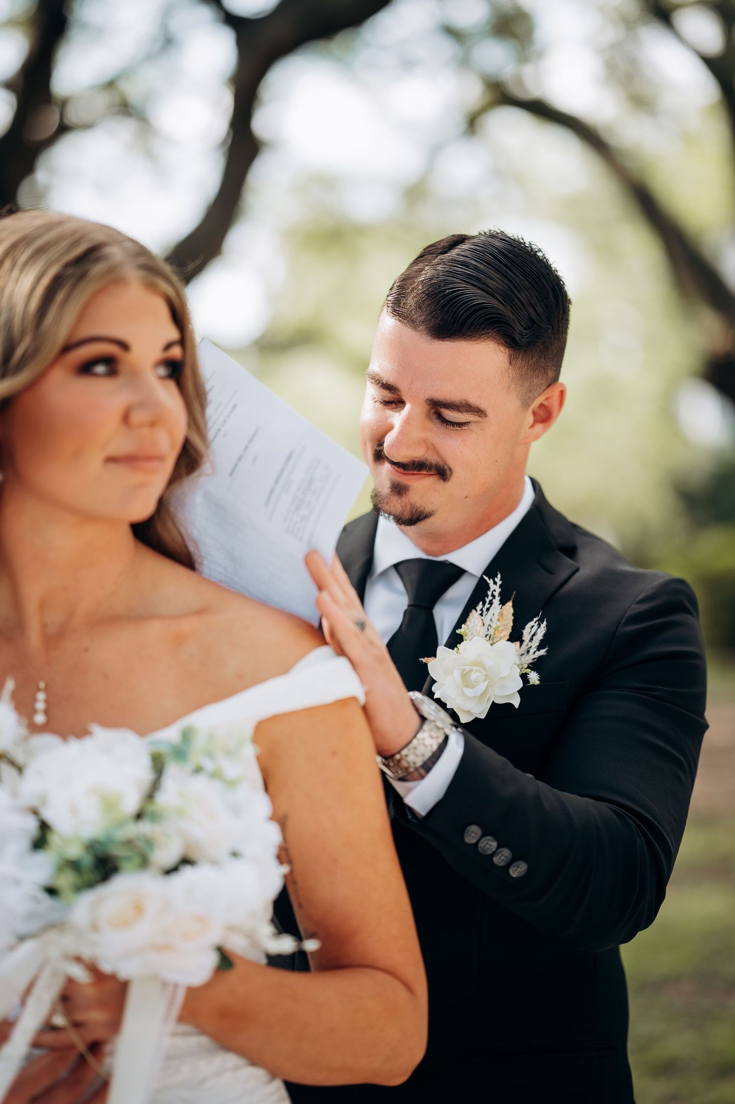 Groom in black suit with white boutonniere reads vows beside bride holding white bouquet at outdoor wedding ceremony.