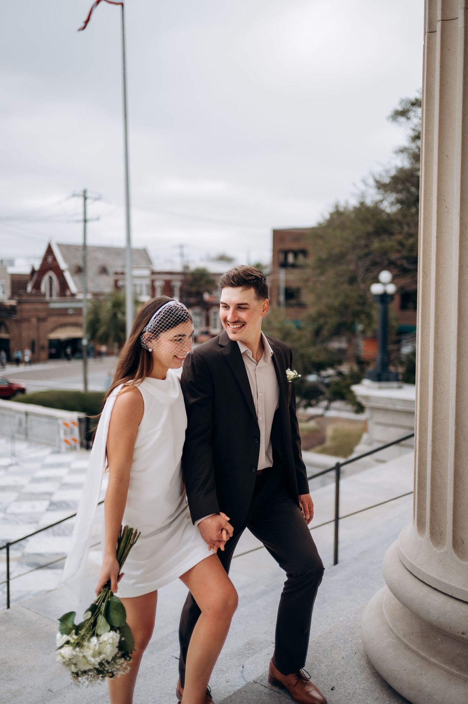 A couple in formal attire shares a joyful moment on steps outside, with the bride holding a white bouquet.