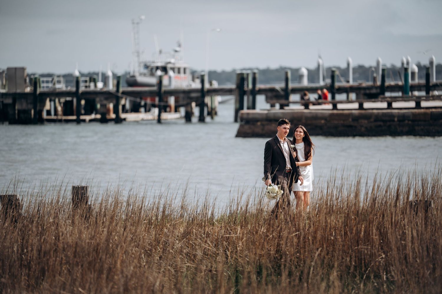 A couple stands together near a marina with tall grass and boats in the background.