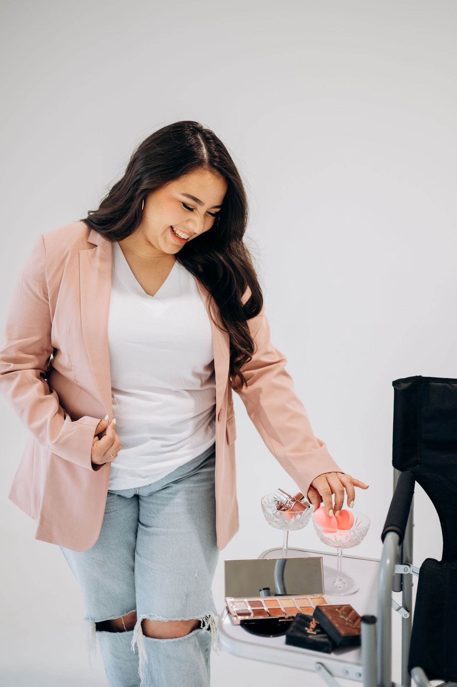 Professional woman in pink blazer and jeans pouring coffee into glass cup at modern white desk setup.