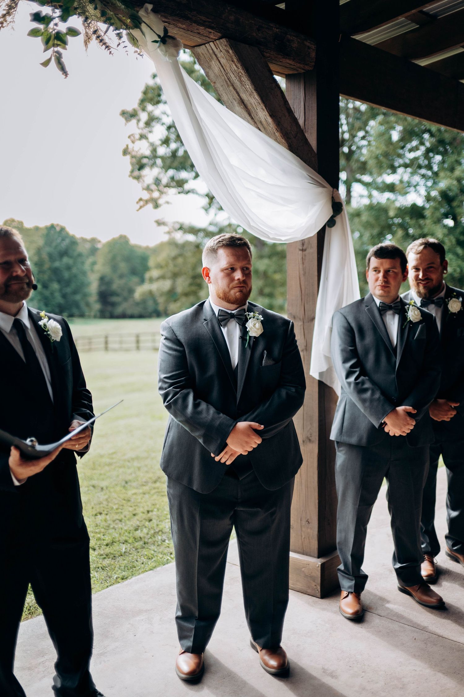 Groomsmen in dark suits lined up under wooden pavilion during wedding ceremony.