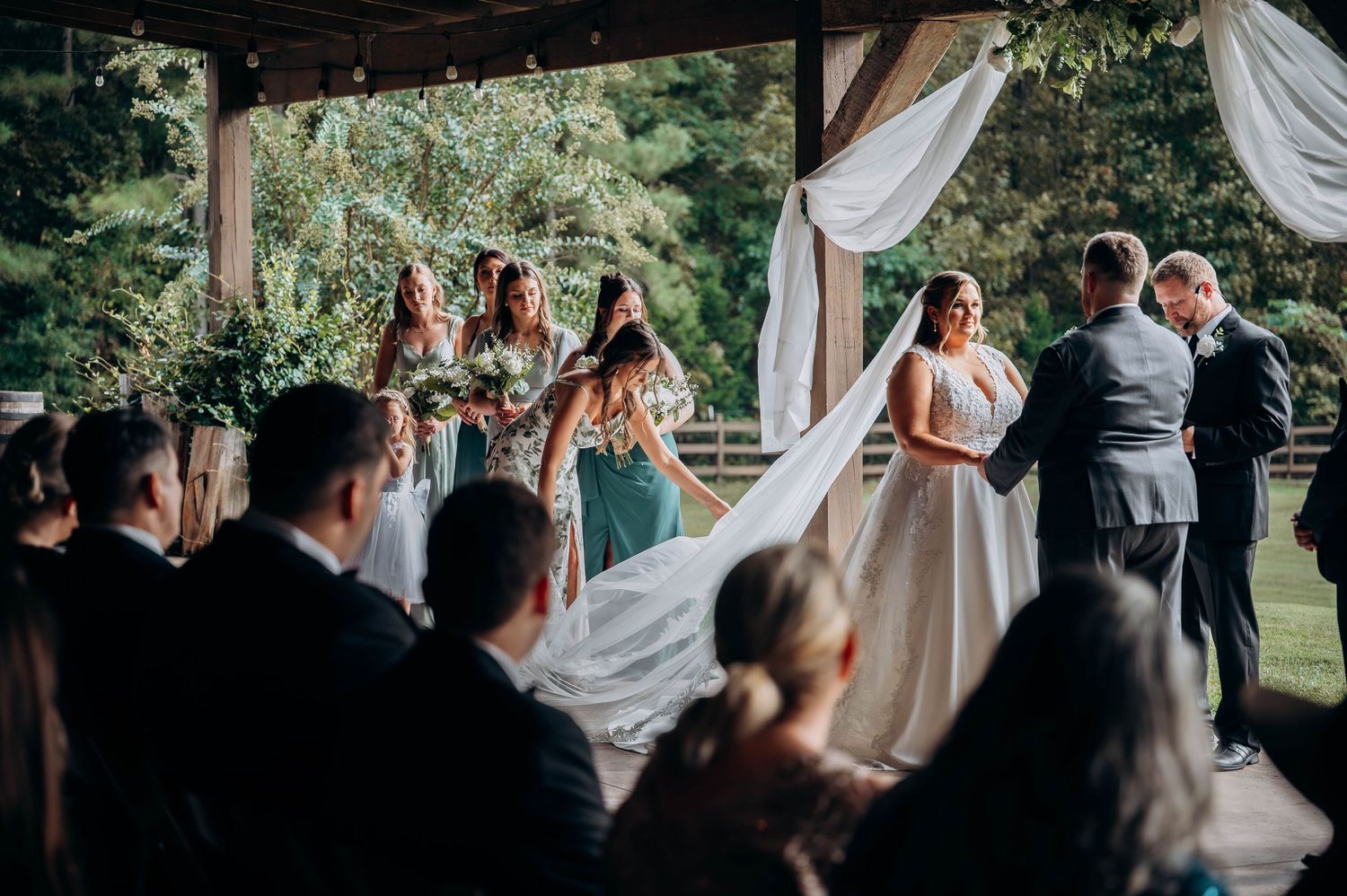 Outdoor wedding ceremony with guests watching under covered pavilion with white drapes.