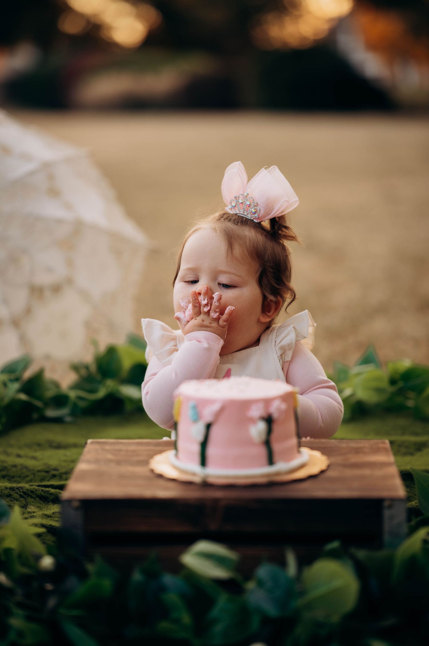 A baby in a party dress sits at a wooden table with a pink birthday cake.