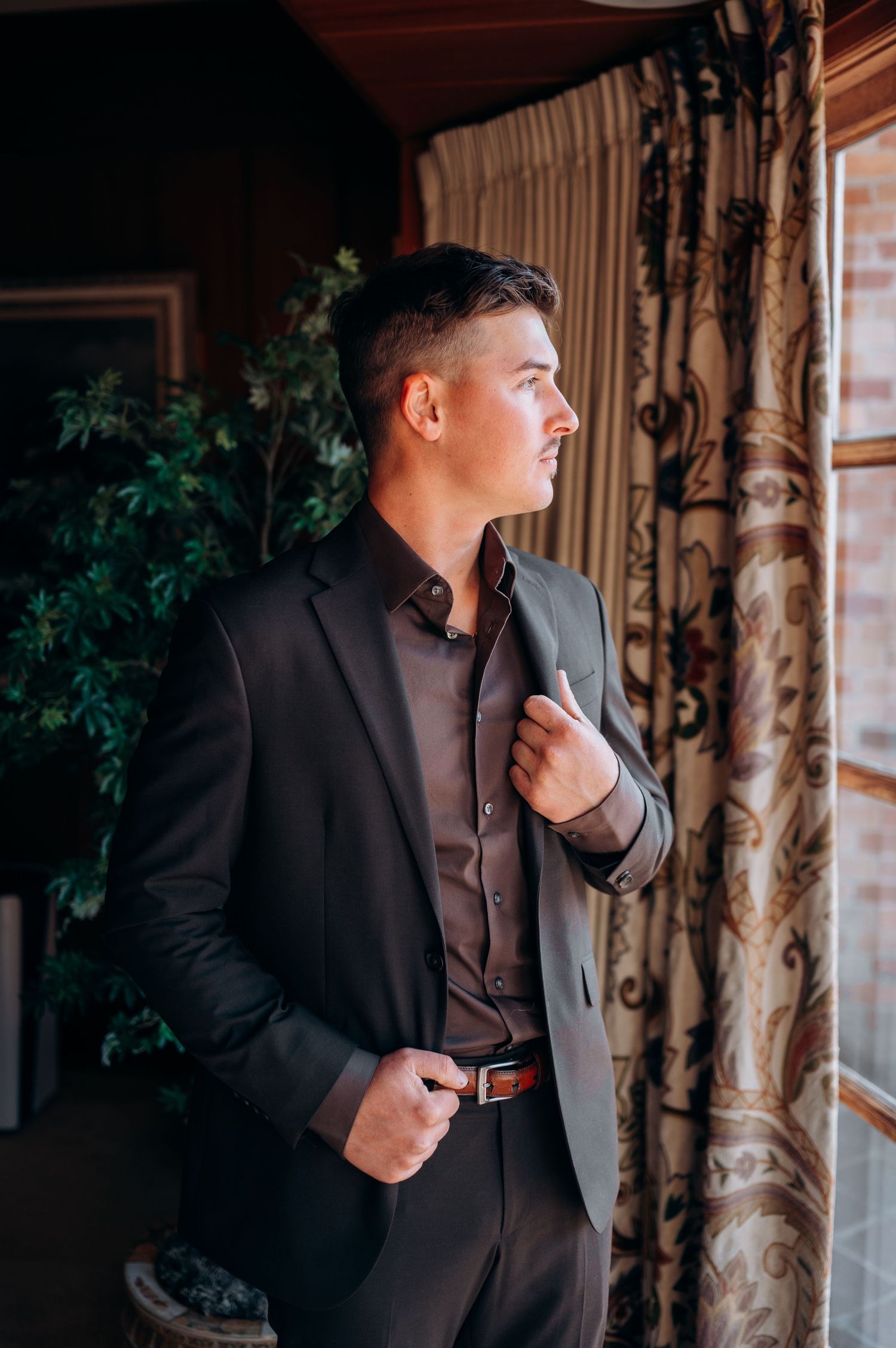 Elegant groom in black suit gazes out window in formal setting with ornate curtains and greenery.