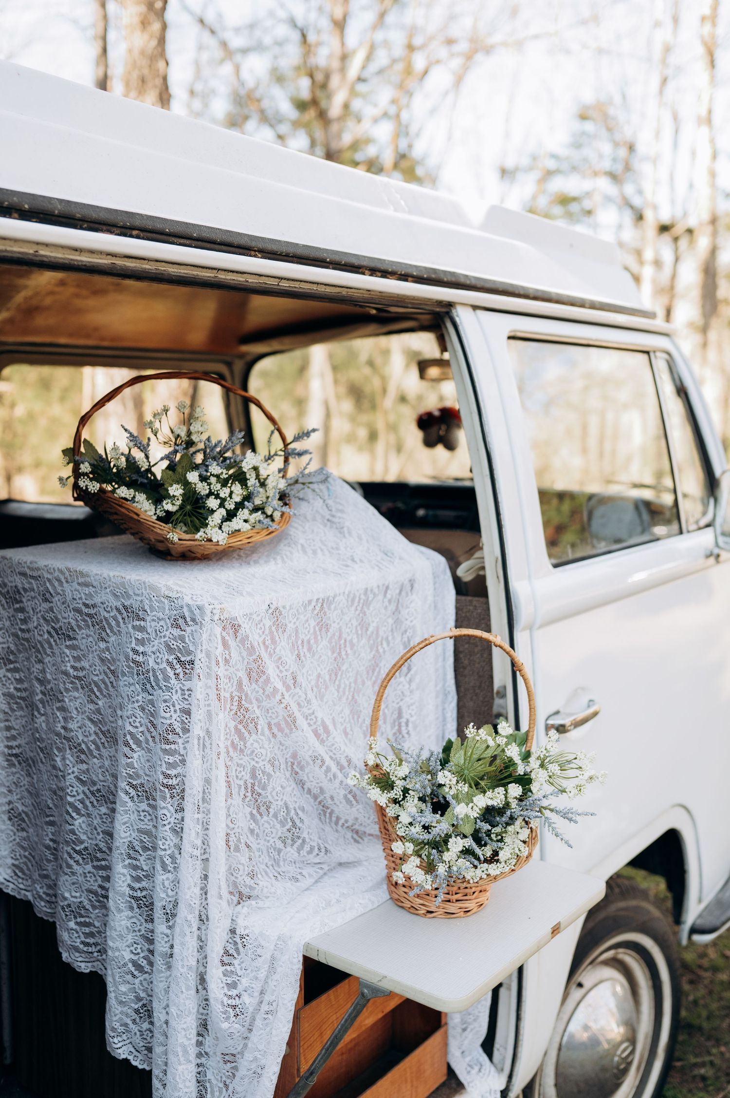 White vintage VW van decorated with lace fabric and floral baskets for a rustic wedding setup.