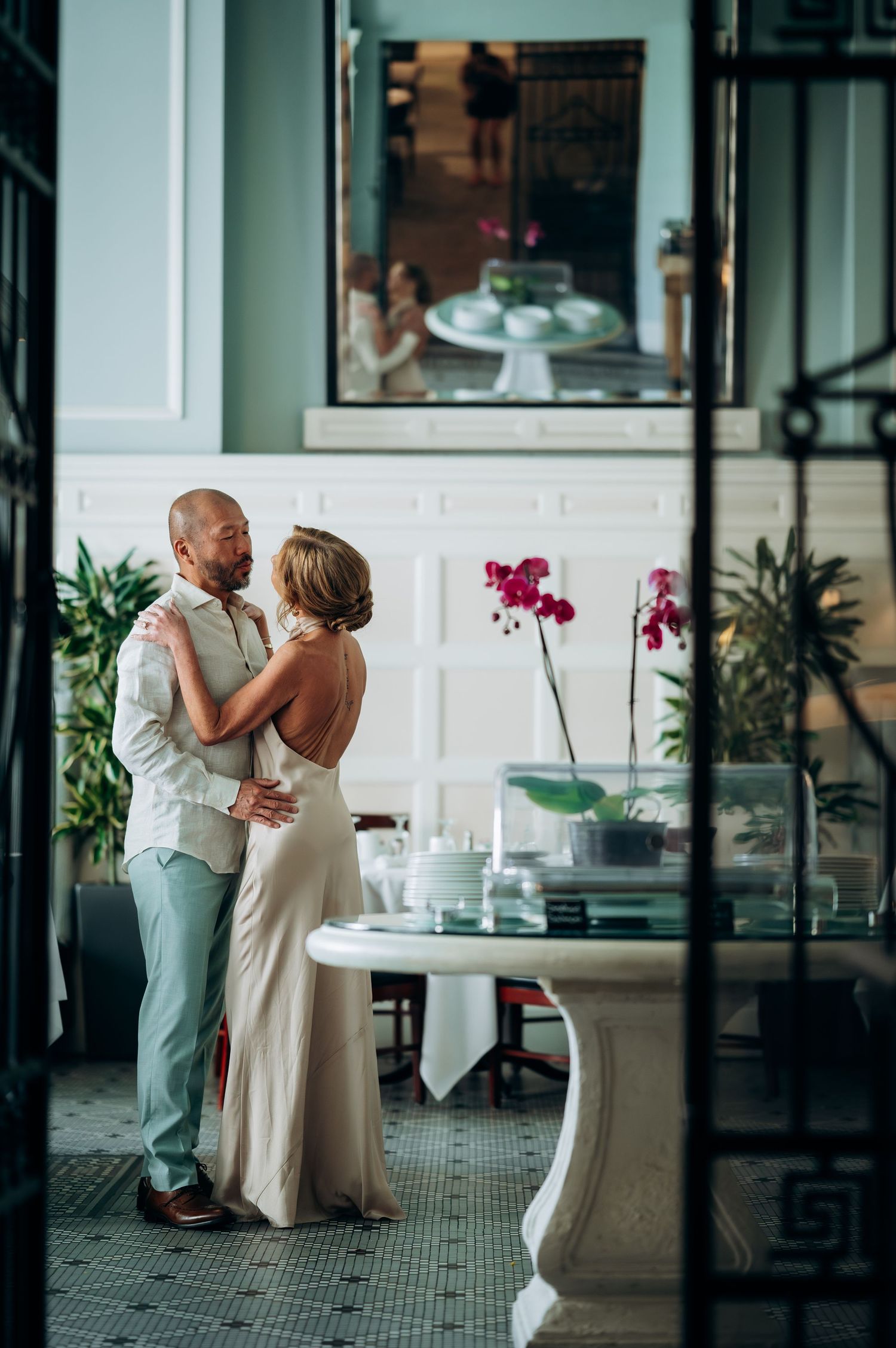 Elegant couple embracing in a luxurious venue with orchids and mirror reflections, wearing wedding attire.