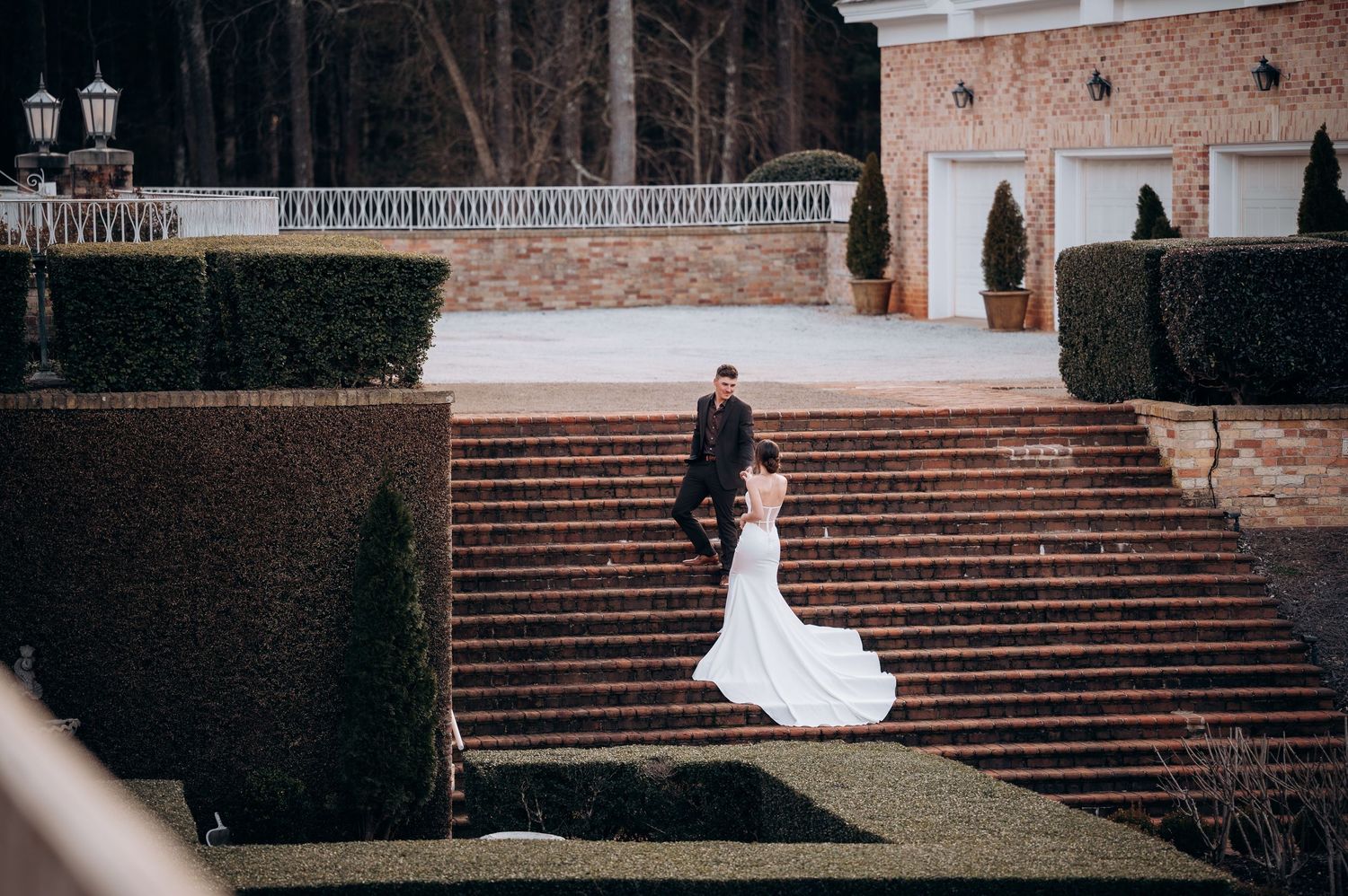 Bride and groom pose on elegant brick staircase surrounded by manicured hedges at a grand wedding venue.