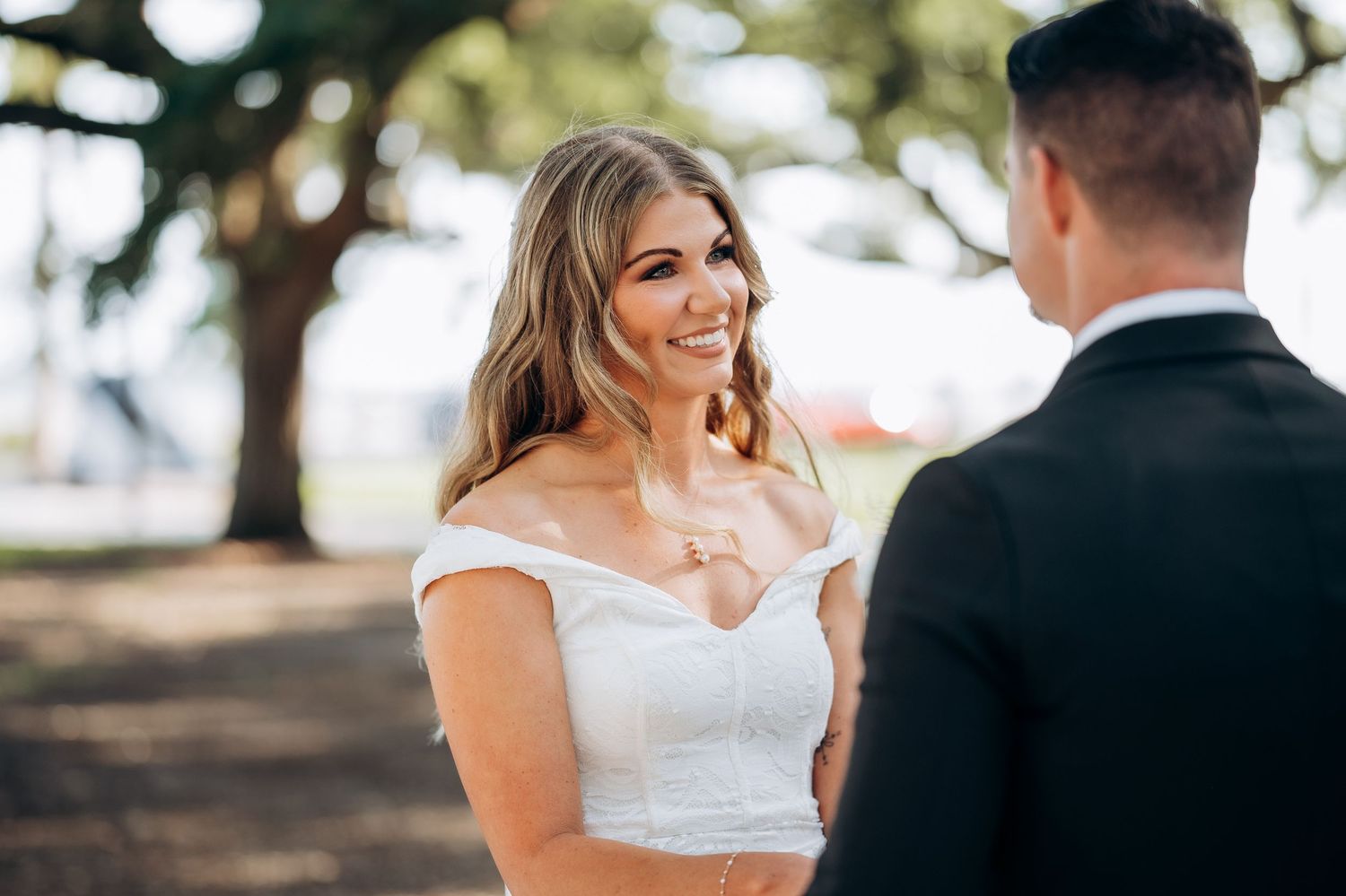 Smiling bride in off-shoulder white wedding dress holding hands with groom in black suit outdoors near trees.