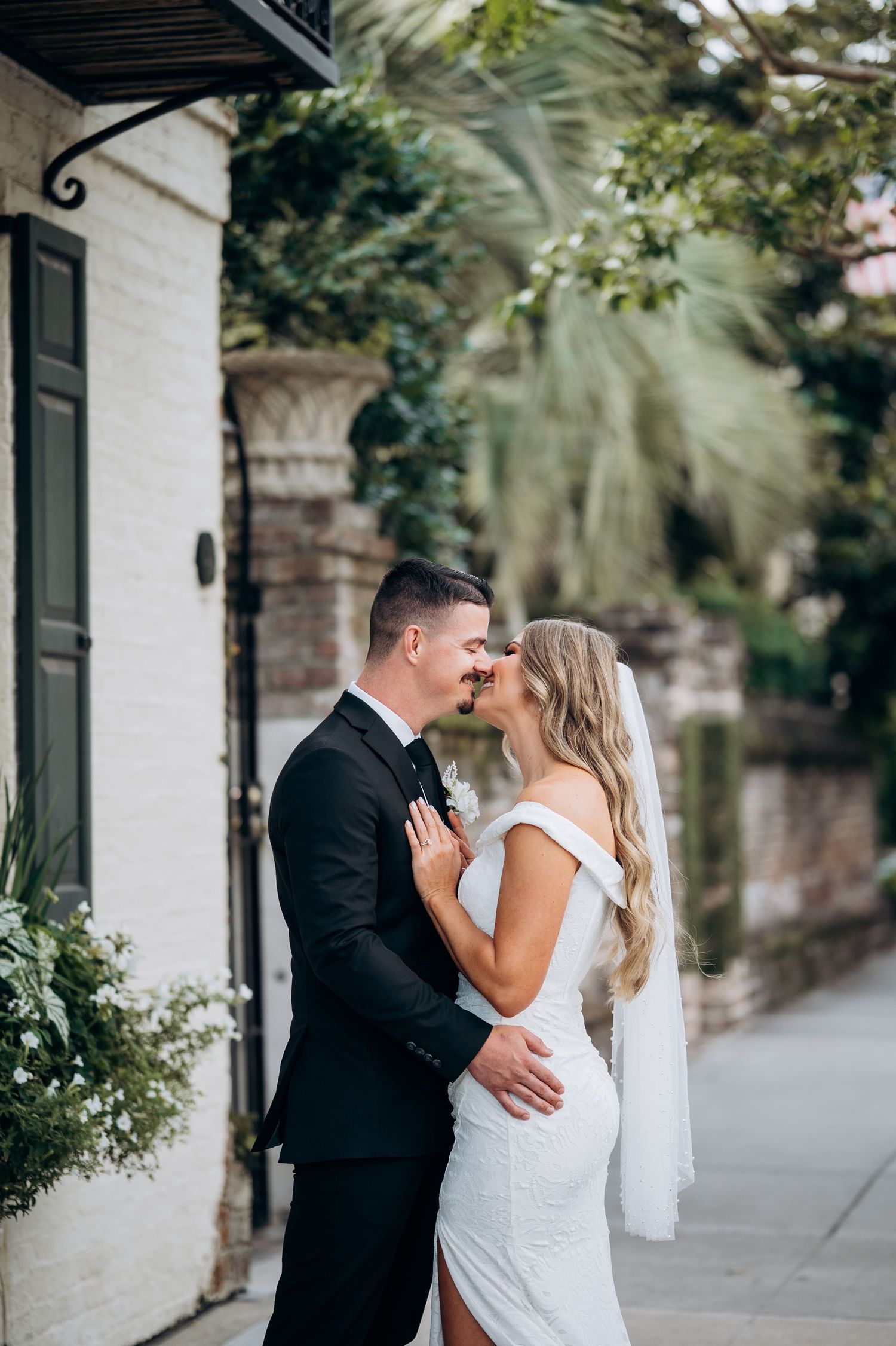 Bride and groom share a kiss on charming Charleston street with white brick building backdrop.
