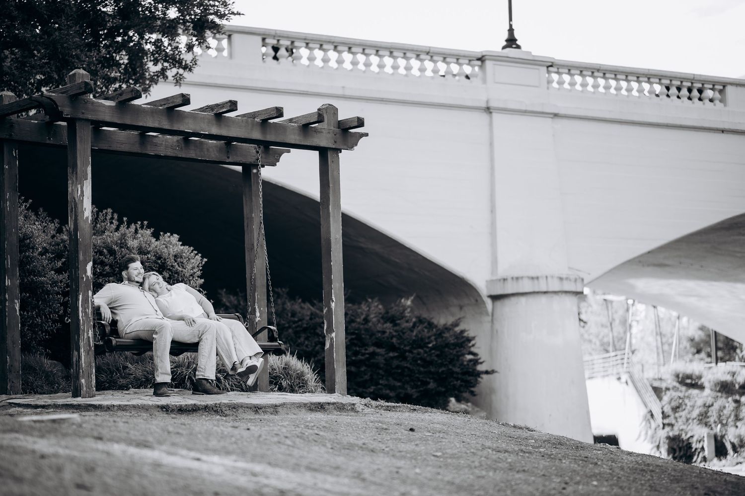 Black and white photo of a couple relaxing on a bench under a wooden pergola near a stone bridge.