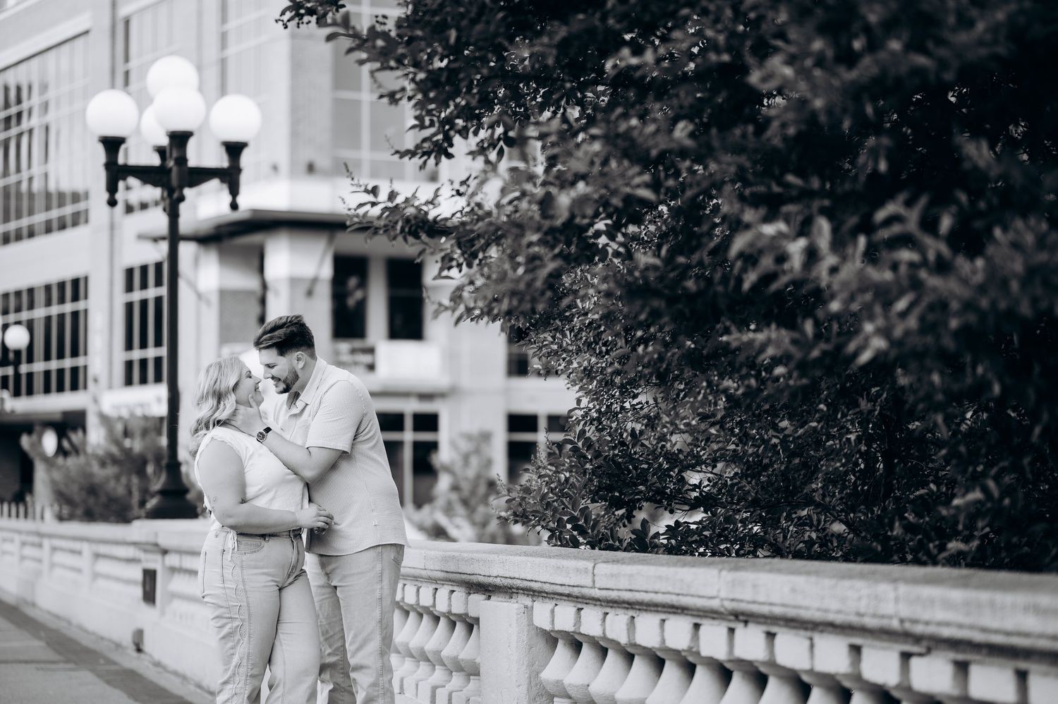 Black and white photo of a romantic couple embracing on a bridge in an urban setting with trees and buildings.