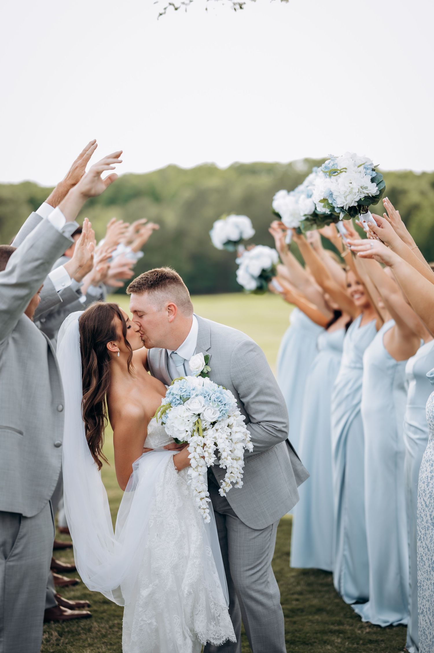Newlyweds share first kiss as wedding party in gray and light blue raises bouquets in celebration.