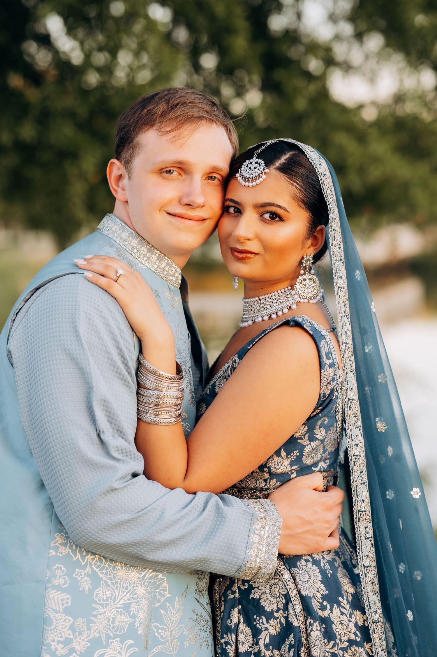 Couple in traditional Indian wedding attire, woman in blue lehenga with jewelry, man in matching sherwani, embracing outdoors.